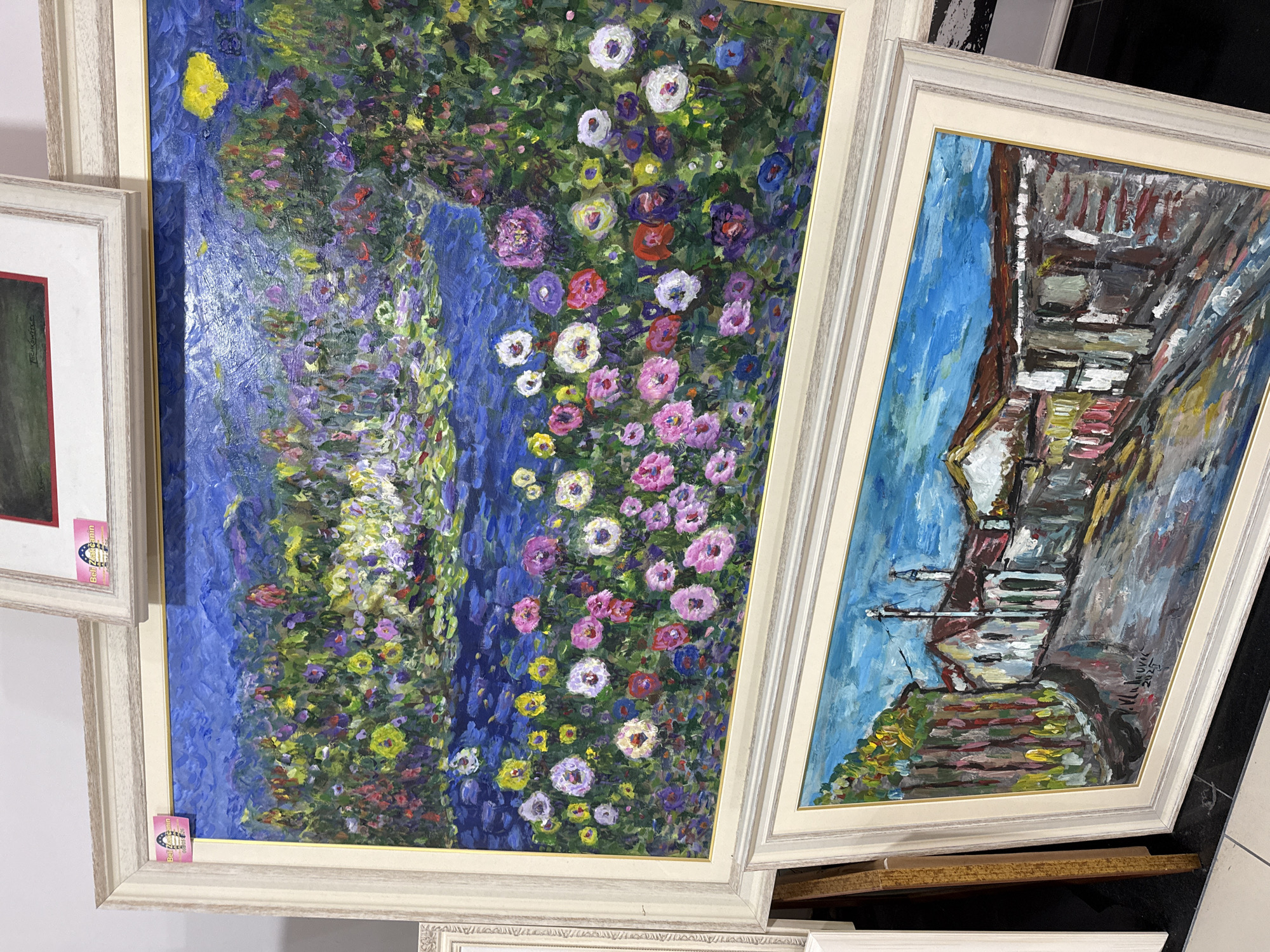 Two framed paintings are displayed. The top painting is a vibrant, impressionistic depiction of a pond surrounded by abundant, colorful flowers in shades of pink, purple, yellow, and red. The water of the pond is a deep blue, reflecting the lush foliage and sky. A single yellow flower or sunburst is visible in the upper right corner. The bottom painting is a more representational, yet still loosely painted, scene of a street in a village. White and brown buildings with red-tiled roofs line a narrow, grey street. There are hints of green trees and foliage on the left side of the street, and a utility pole with wires is visible against a bright blue sky. The bottom painting is signed "V. MLIKOVIC 2025" in the lower right corner. Both paintings are framed in light-colored, ornate frames. The paintings are stacked or leaning against each other, suggesting they are in an art gallery or exhibition space. There are pink stickers with a horseshoe and the word "Beli Zajcanin" on the frames of both paintings.
