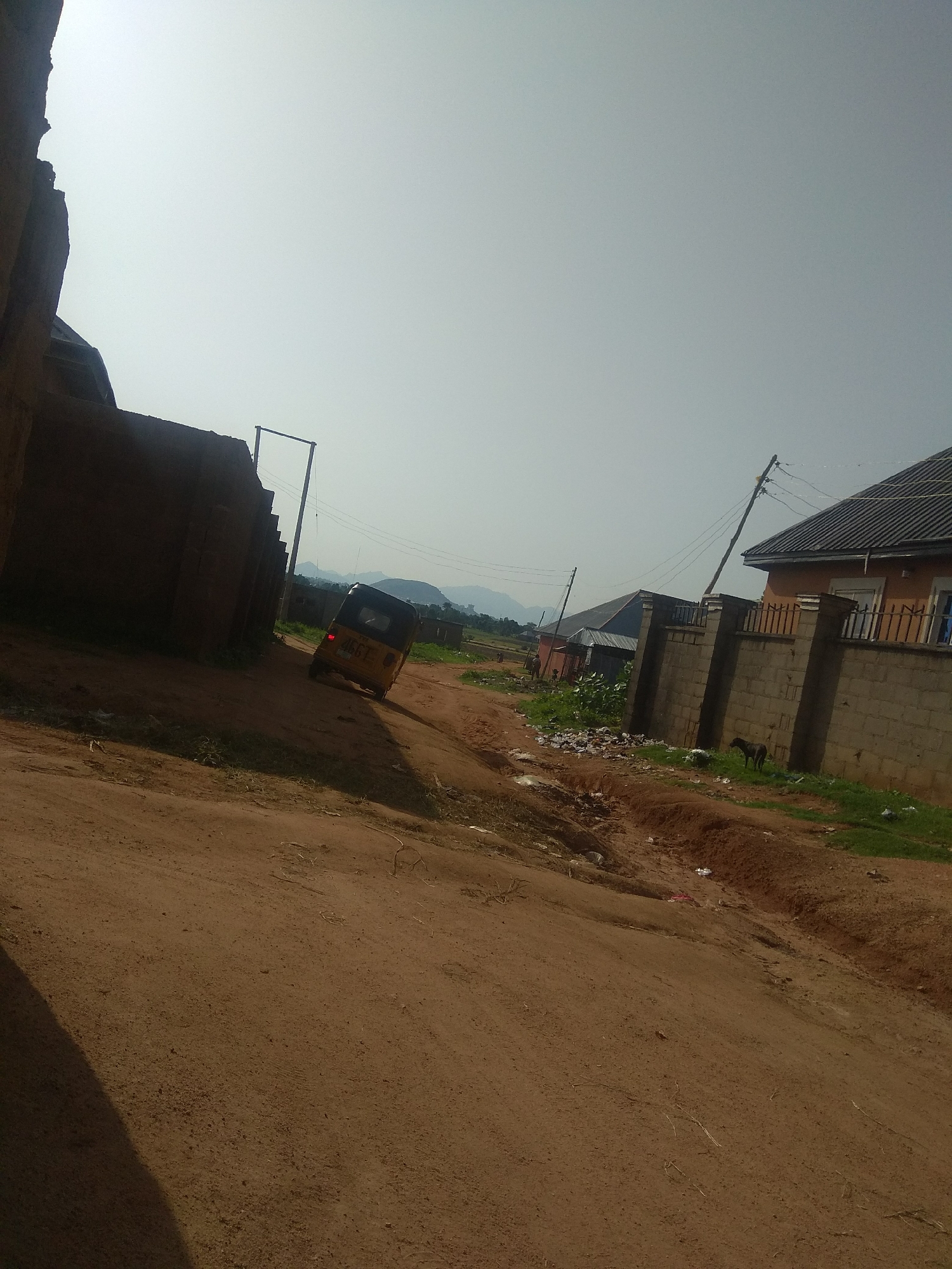 The image depicts a dirt road in a residential area of Jalingo, Nigeria, under a bright, hazy sky. A yellow and black motorized tricycle, commonly known as an "okada" or "keke napep," is parked on the side of the road, facing away from the viewer. To the left of the road, a dark, unfinished brick wall dominates the foreground, casting a shadow across the dirt. Across the road, several houses with corrugated iron roofs and stone-block walls are visible. In the middle distance, a small dark dog stands near a brick wall with a metal railing. The environment appears somewhat unkempt, with scattered debris along the roadside and a shallow trench or erosion gully visible in the foreground. Distant hills or mountains are faintly visible on the horizon. The scene suggests a typical urban or peri-urban setting in Nigeria, with the presence of local transportation, residential structures, and unpaved roads.
