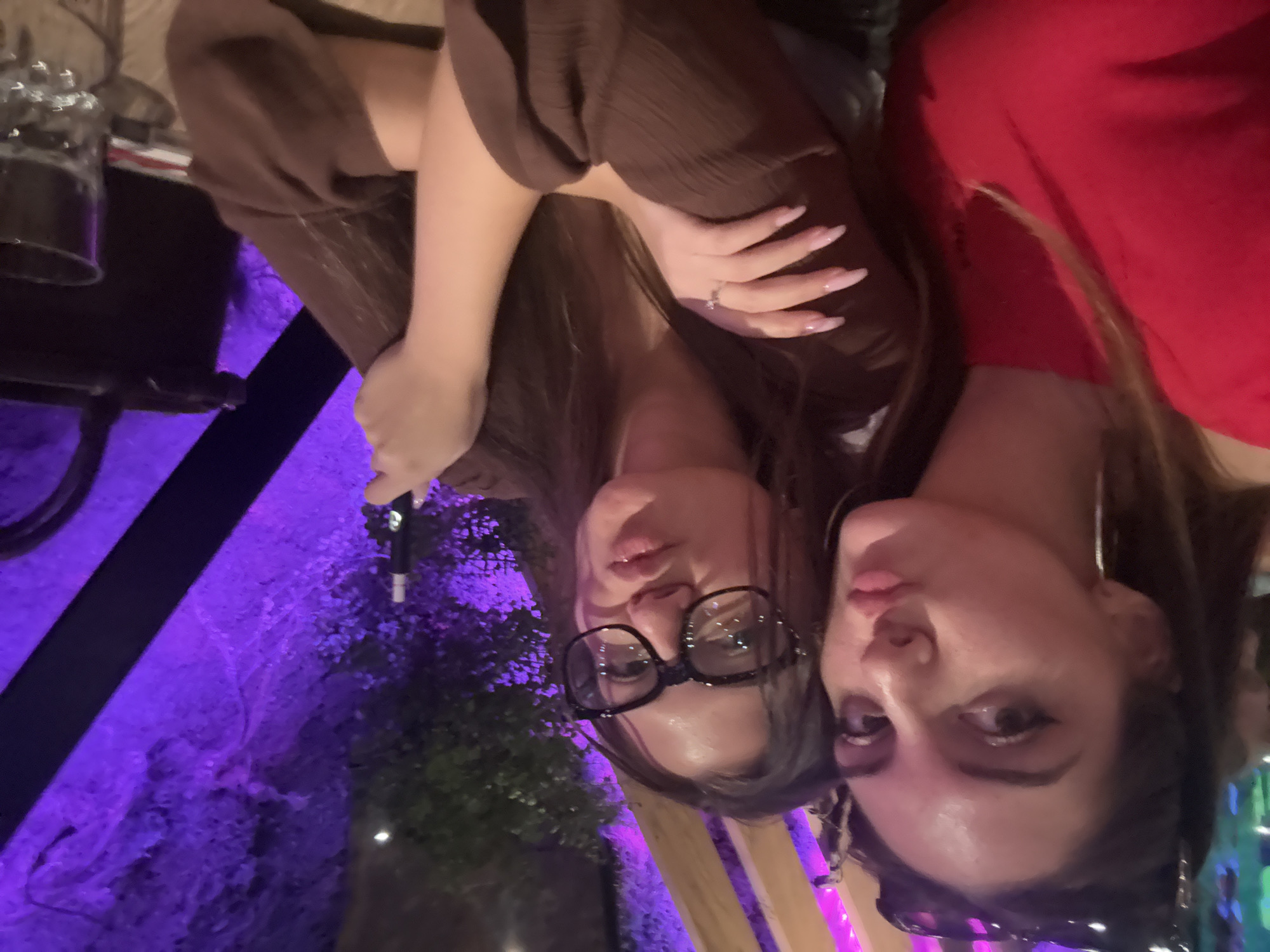 Two women are posing for a photo. The woman on the left is wearing glasses and a brown top. The woman on the right is wearing a red top and hoop earrings. They appear to be at an outdoor restaurant or bar with purple lighting. The location might be in the city of Sofia, Bulgaria.