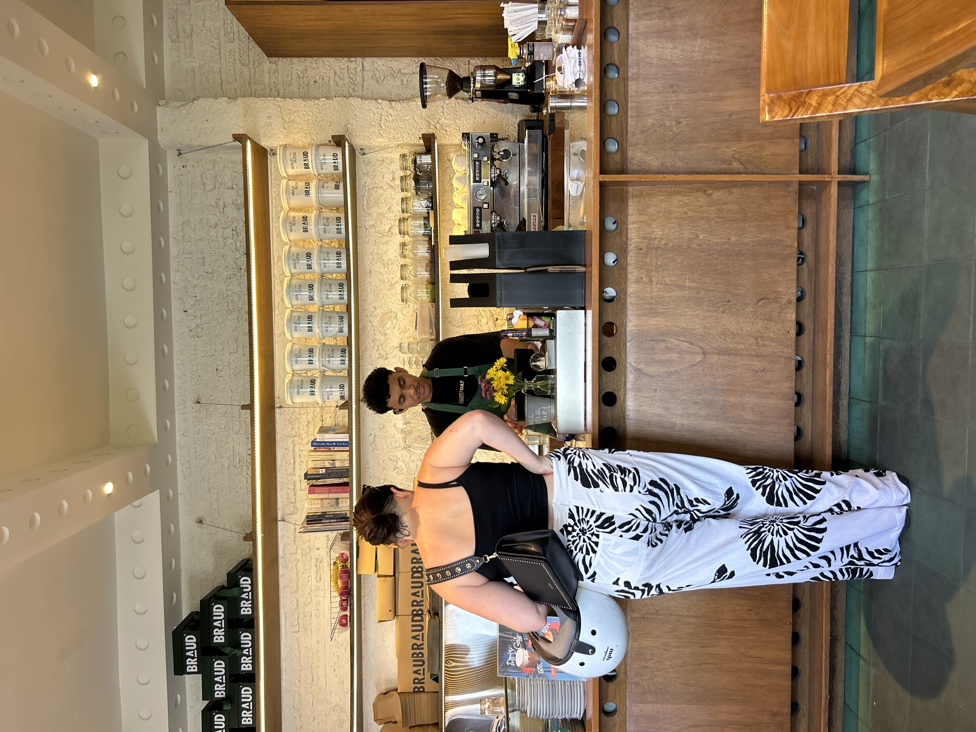 A woman with dark hair pulled up stands at a counter in a coffee shop in North Kuta, Indonesia. She is wearing a black tank top and white pants with a black palm leaf pattern. A black purse with gold stud detailing is slung over her left shoulder, and a white helmet is tucked under her right arm. She is facing away from the camera, looking at a barista. The barista, a young man with dark curly hair, wears a green apron and black shirt. He is standing behind the counter, interacting with the woman.
The coffee shop counter is made of dark wood, with shelves above displaying various items. The shelves hold stacks of green boxes with the word "BRAUD" printed on them, a basket of red and yellow items, several books, and rows of white containers also labeled "BRAUD." To the right of the counter, a commercial espresso machine and a coffee grinder are visible. Jars filled with what appear to be ingredients are also on display on a higher shelf. The walls behind the counter are made of white brick. The floor is tiled in a dark green color. The lighting in the shop is bright, with a series of circular lights recessed into the ceiling. The overall atmosphere is that of a modern, stylish coffee shop.