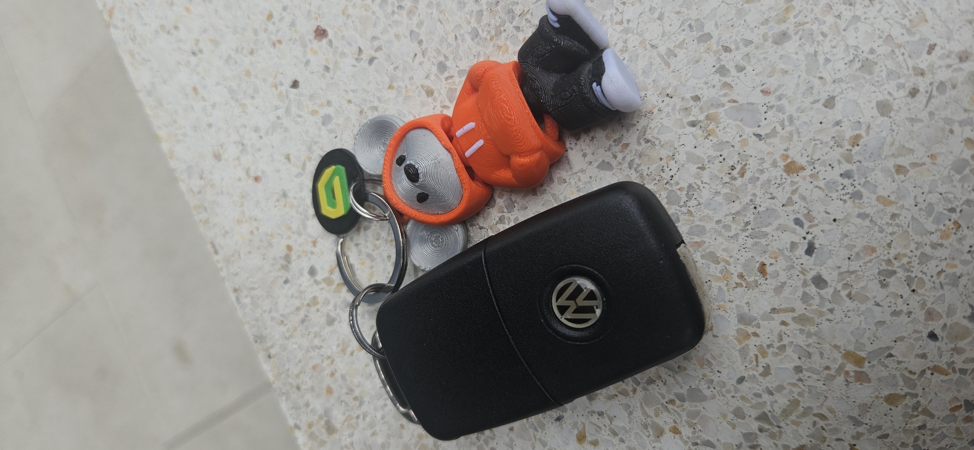 The image presents a set of car keys and associated keychains resting on a light-colored, speckled surface, likely terrazzo or a similar composite material. A smoother, lighter surface is partially visible on the left, forming a corner with the speckled background.
The primary object is a black Volkswagen (VW) car key fob, featuring the distinctive silver VW logo centrally placed. One end of the key fob appears worn. Attached to the fob by several silver metal rings are two additional keychains. One is an orange and grey figurine, possibly a character or an animal in a hoodie, with a textured finish suggesting it might be 3D-printed. It has a grey face with small black eyes and an orange body adorned with white markings. Its visible limbs are black with white tips. The second keychain is a flat, black oval shape with a stylized green and yellow letter 'D' emblem.
The scene is illuminated by soft, diffused light, indicating daytime. No specific activity is occurring; the objects are static. There are no people visible, nor are there any discernible emotional cues. The textural details of the keychains and the granular pattern of the surface are clearly visible. While the image itself does not contain explicit geographical markers, this media file is situated in the context of Plovdiv, Bulgaria.