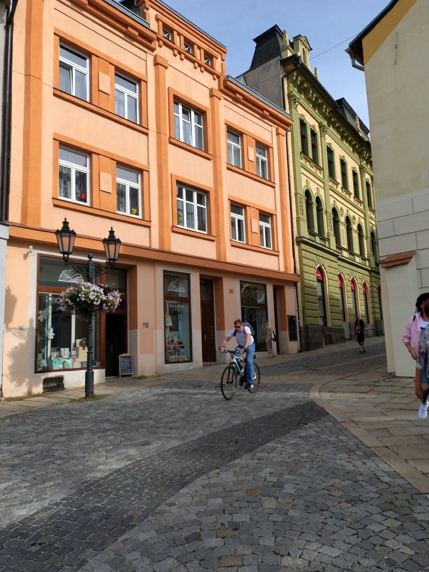 The image captures a street scene in Písek, Czechia. A man on a bicycle rides down a cobblestone street lined with colorful buildings. The building on the left is painted in shades of orange and features multiple windows. The building in the middle ground is painted green with ornate architectural details. To the right, a portion of a light-colored building is visible. A lamppost with hanging flowers stands in front of a storefront on the orange building, which appears to be a bookshop with books displayed in the window. Another person is visible in the background, walking away from the camera. The sky is clear and blue.