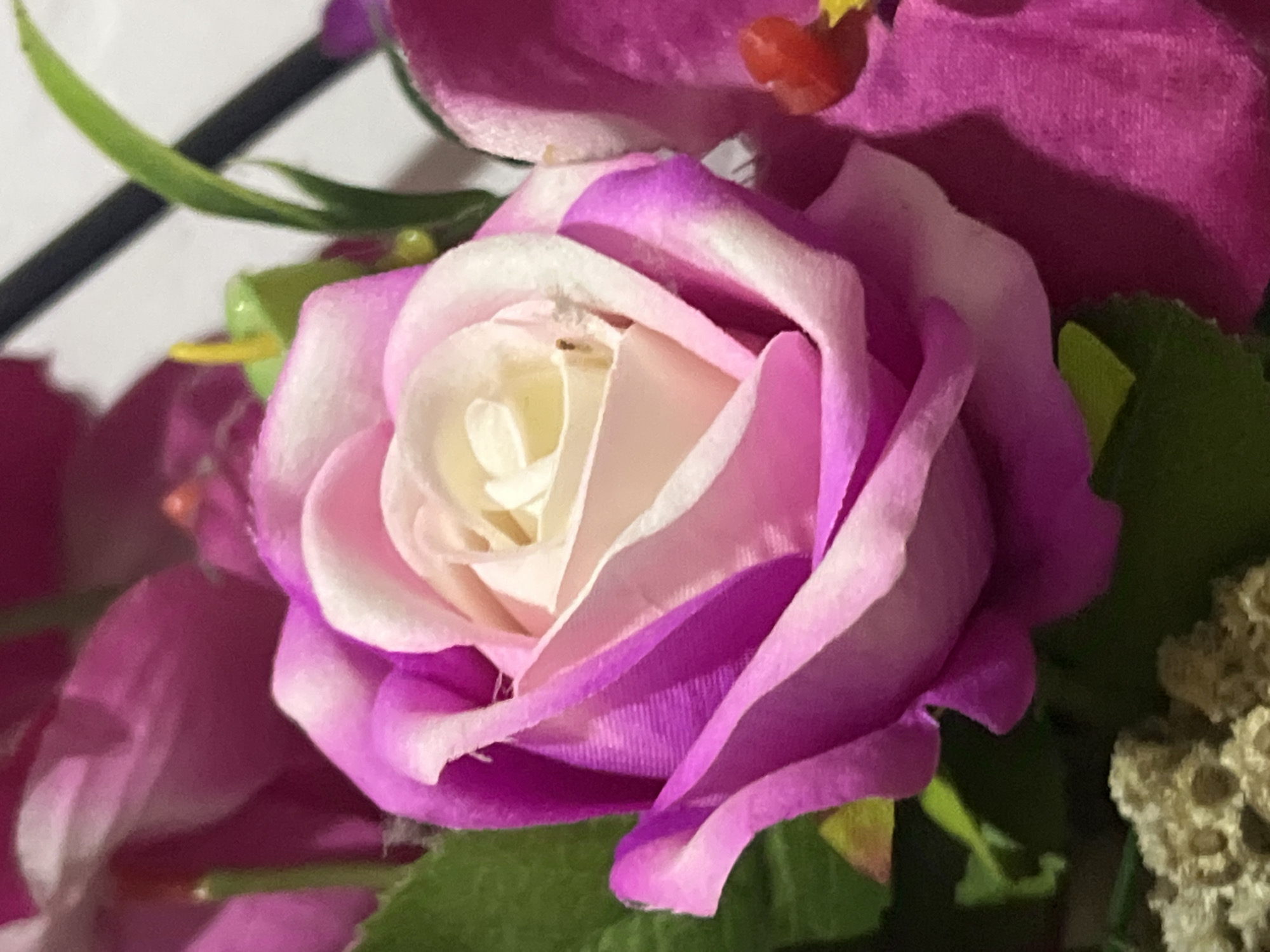 The image is a close-up, vertical, eye-level shot of artificial flowers. The primary subject is a pink and white rose with layered petals, featuring a bright yellow stamen in the center. To the right of the rose, a vibrant fuchsia orchid is visible, adorned with a small red bead-like detail. Green leaves and other floral elements in shades of pink and fuchsia are present in the background, some of which are out of focus. The lighting suggests an indoor setting with a diffused light source. The image does not depict any recognizable urban elements or locations within Denpasar, Indonesia.
