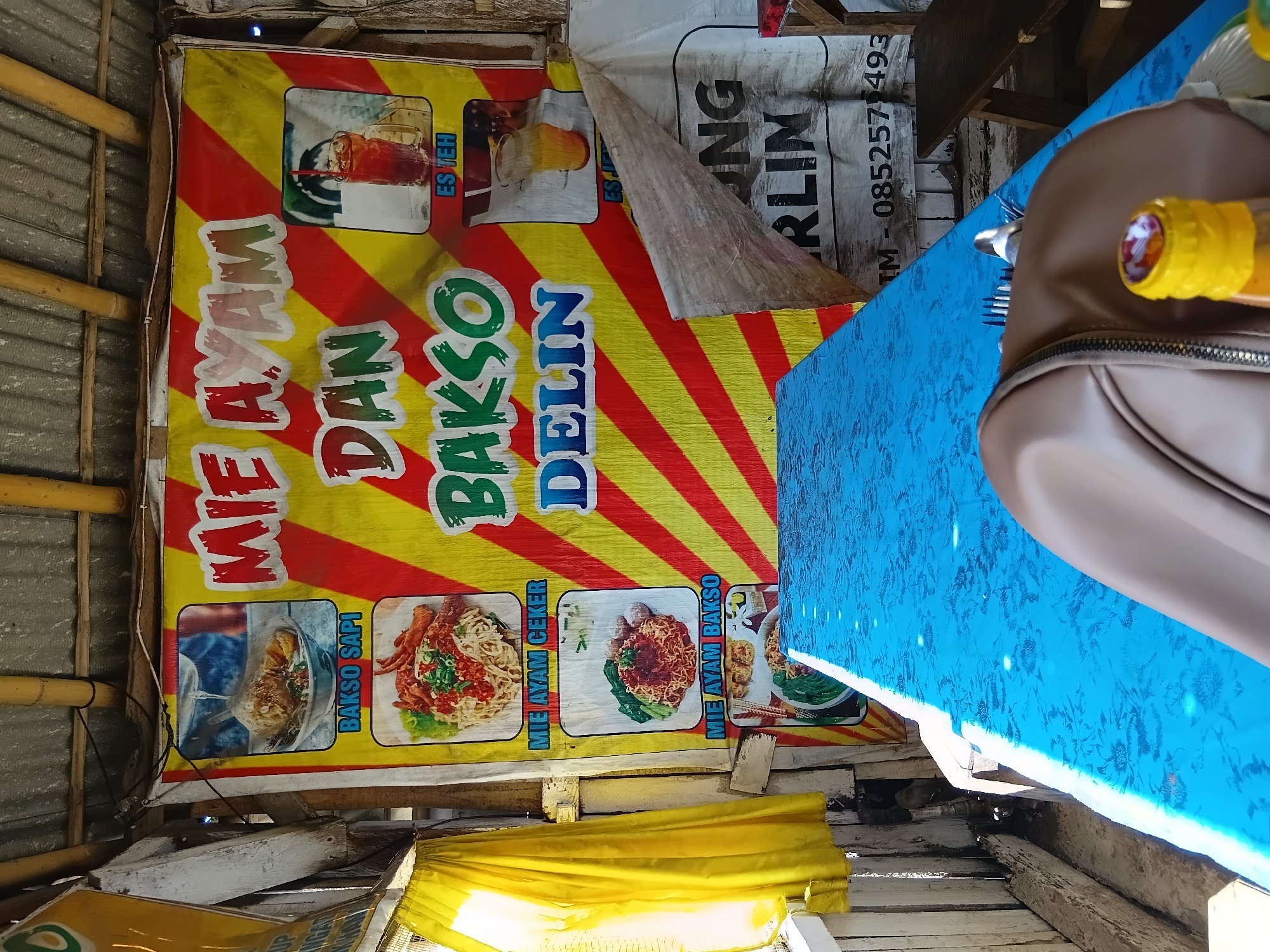 An outdoor food stall in Kasiyan Timur, Indonesia displays a large banner advertising "MIE AYAM DAN BAKSO DELIN." The banner features images of noodle dishes and drinks, with bold red and yellow diagonal stripes. To the left, a yellow curtain hangs, partially obscuring a wooden structure. In front of the banner, a blue tablecloth covers a table. On the table, a brown bag and a partially visible bottle with a yellow cap are present. A white tarp with black lettering, including "UNG RLIN" and a phone number, is partially visible to the right of the banner. The overall environment suggests a simple, open-air eatery.