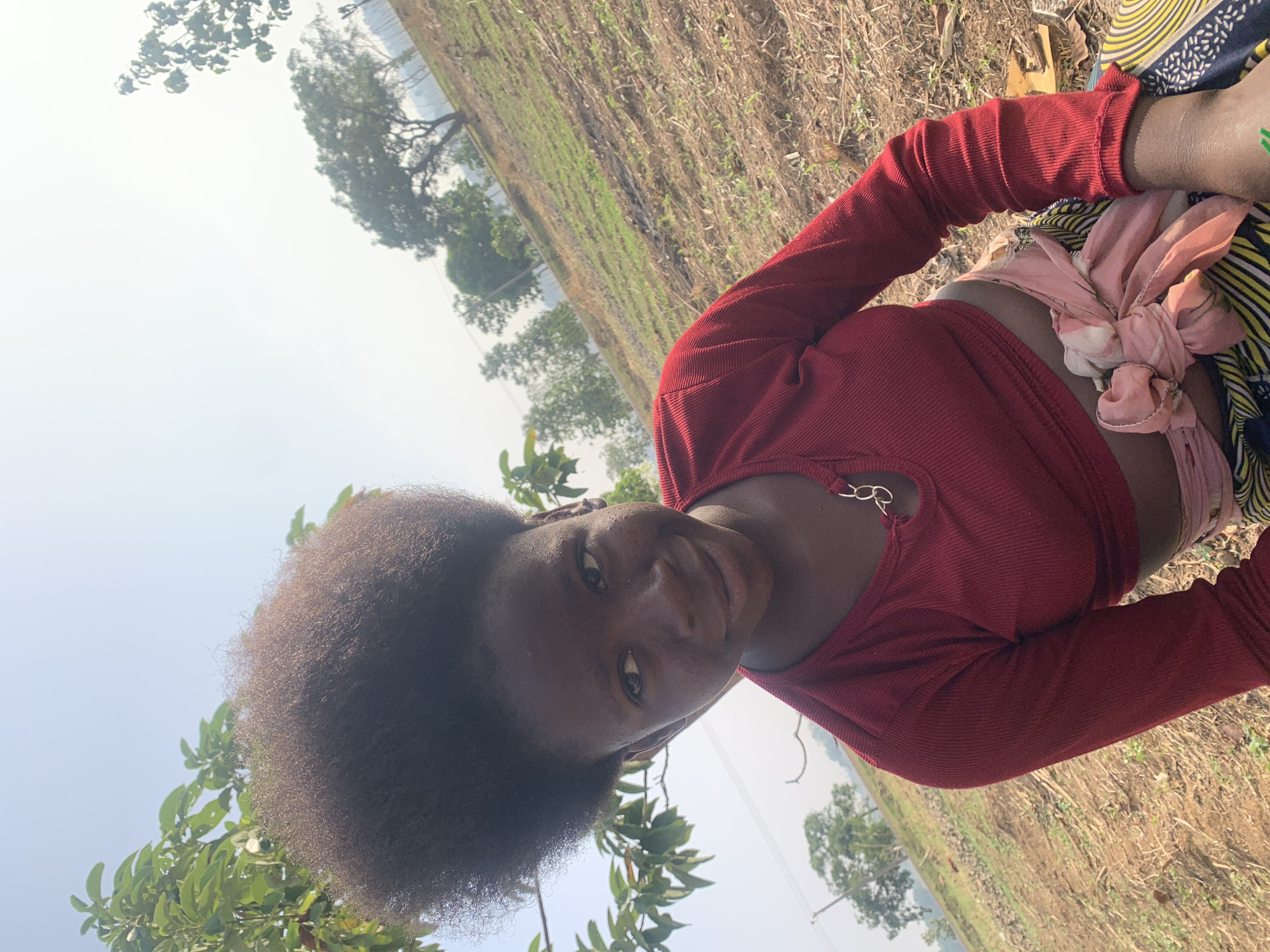 An outdoor portrait captures a young Black woman in what appears to be Agban, Nigeria. The image is rotated 90 degrees counter-clockwise. The woman, with dark, curly hair, looks directly at the camera with a calm, friendly expression and a slight smile. She is wearing a form-fitting, dark red, long-sleeved ribbed top featuring a cut-out detail on the neckline adorned with a gold chain. Around her midsection, a light pink fabric is tied over a patterned garment, likely a wrapper or skirt, visible in yellow, blue, and black.
The setting is an open, sunlit area, possibly a field or undeveloped land. The background features dry, brown soil mixed with patches of green vegetation. Several trees with green foliage are visible further back, under a bright, hazy sky, suggesting it is daytime, possibly mid-morning or afternoon. The woman appears to be leaning forward or crouching slightly. No specific activity is taking place beyond posing for the photograph. There is no visible text in the image.