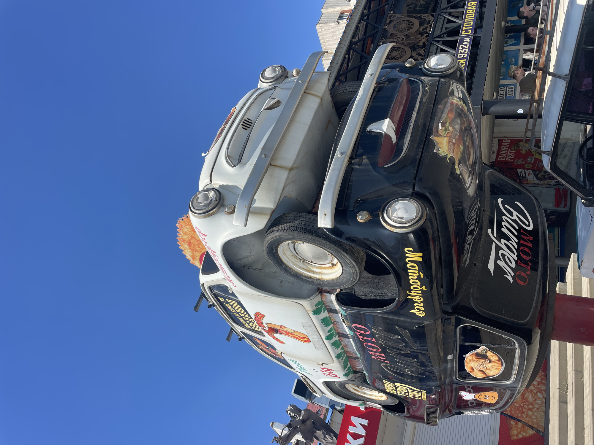 The image shows a roadside attraction in Kamensk-Shakhtinsky, Russia, featuring two vintage cars stacked vertically. The top car, a white sedan, is adorned with graphics of a hot dog and green festive foliage along its side. A large orange, fan-like decoration is visible on its roof. The bottom car, a black sedan, is positioned upside down and displays "MOTO Burger" in stylized script on its side. It also features a sticker of a hamburger on its underside. Both vehicles are mounted on a red pole. In the background, a building with a multi-story structure and an awning is visible, along with people walking below. A sign with Cyrillic lettering and "932 KM" is also present. The sky is clear and blue.