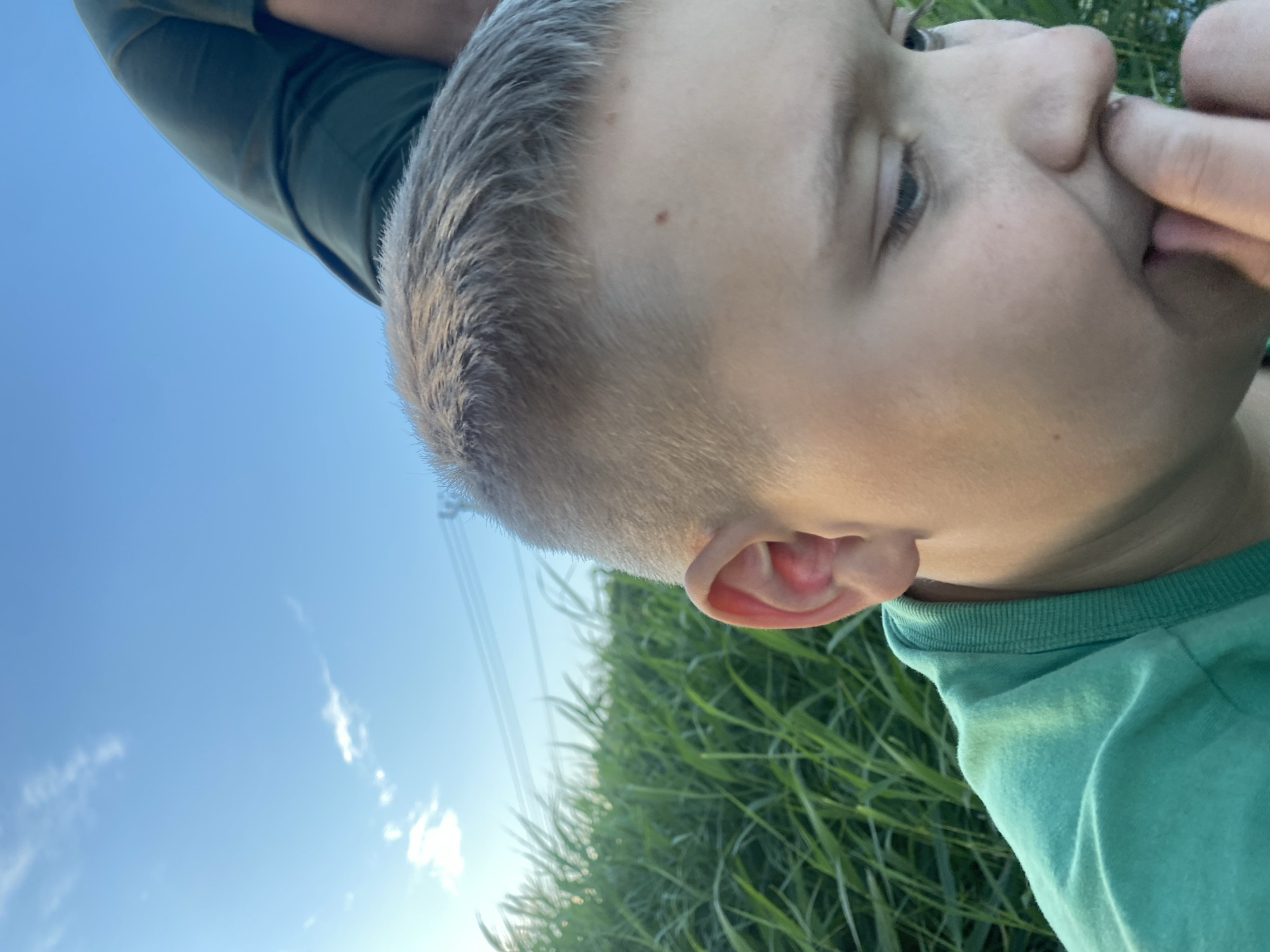 This is a close-up, low-angle, outdoor shot of a young boy's profile. He has short, light brown hair and is wearing a teal t-shirt. His right hand is near his face, with a finger touching his nose. The background is a bright blue sky with some wispy white clouds, and a field of tall green grass. The lighting suggests it is daytime, likely late afternoon or early evening, given the warm light. The overall mood appears relaxed and natural, possibly capturing a candid moment outdoors. There are no visible objects or text in the image. The location context provided is Афанасьево, Russia.