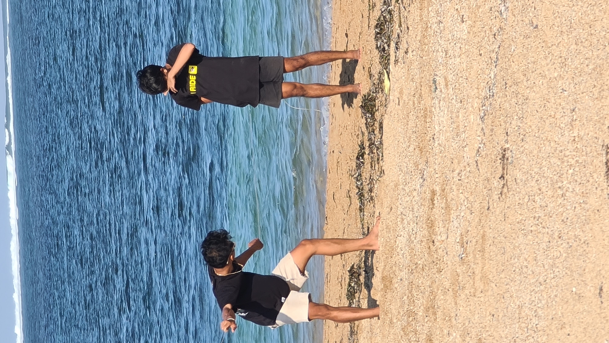 In Denpasar, Indonesia, two young men are on a sandy beach. The man on the left, wearing a black t-shirt and light-colored shorts, is bent over with his hands near his knees, appearing to be stretching or performing a physical activity. The man on the right, in a black t-shirt and dark shorts, is standing and urinating, with a stream of urine visible on his legs. The background features the blue ocean with waves and the horizon. The beach sand is light brown with scattered seaweed.