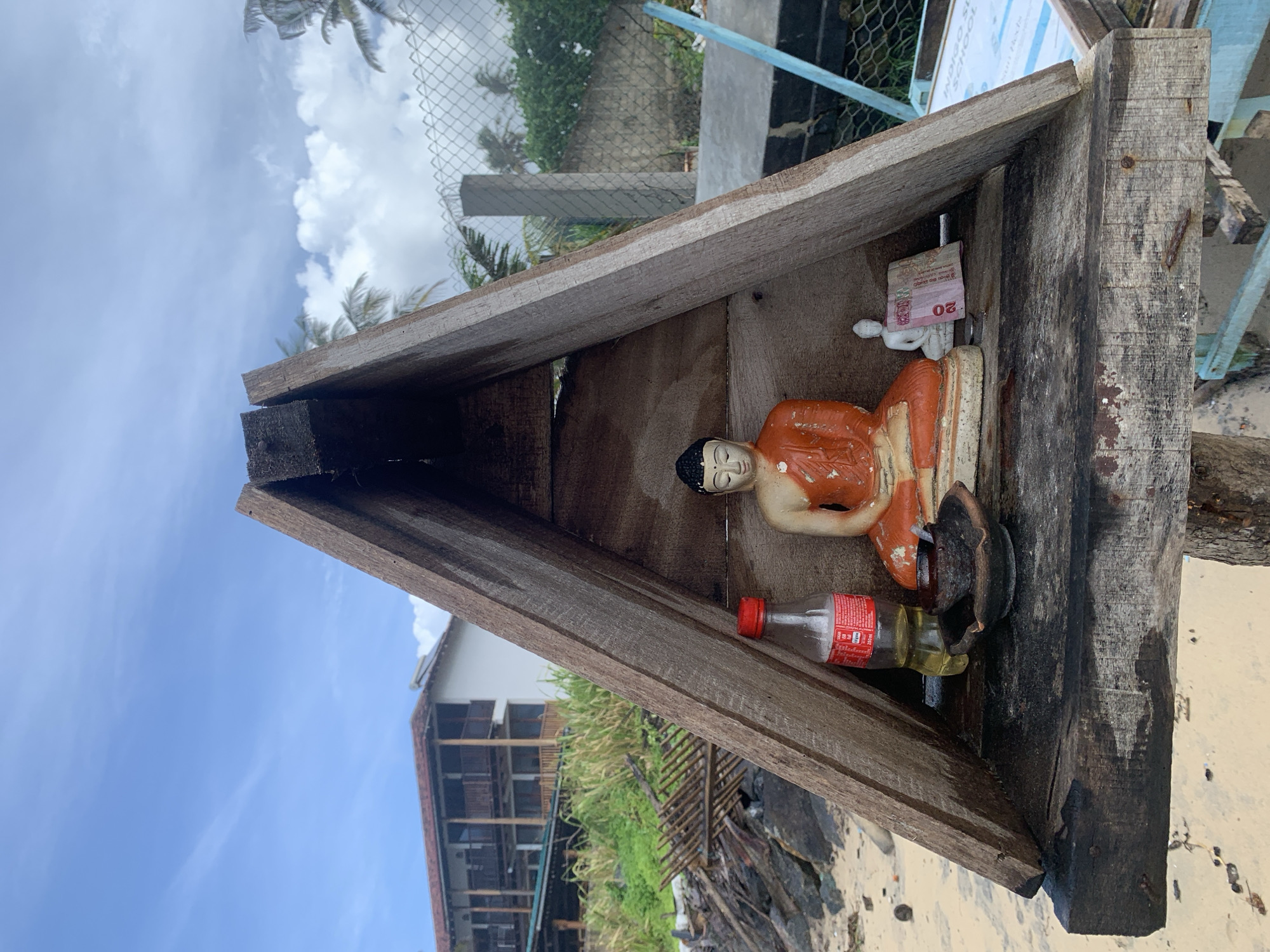 In Kathaluwa, Sri Lanka, a small, weathered wooden structure, triangular in shape, stands on a sandy beach. Inside this structure, a shelf holds several items. A seated Buddha statue, vibrantly colored in orange robes, is the central figure. To its left, a clear plastic bottle with a red cap, appearing to contain a yellow liquid, rests beside a small, dark ceramic dish. To the right of the Buddha statue, a miniature white Buddha figure sits, with a pink banknote tucked behind it. The background reveals a cloudy sky with palm trees, a chain-link fence, and a multi-story building with balconies. The surrounding environment includes rocks and vegetation, suggesting a coastal setting.
