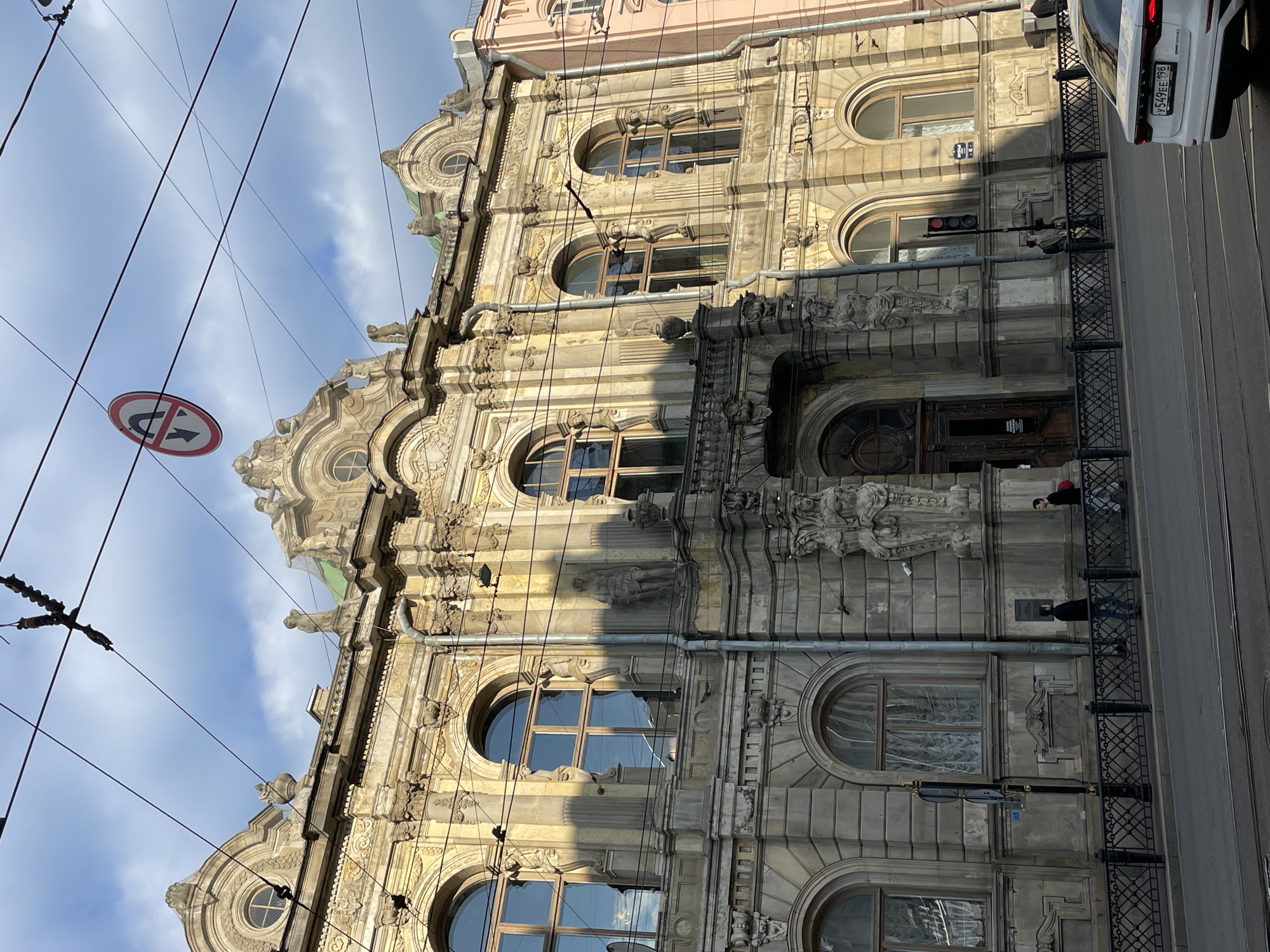 The image shows a tall, ornate building in Saint Petersburg, Russia, likely a historical or cultural landmark, possibly the Elisseeff Emporium. The building features a heavily decorated facade with arched windows, elaborate stonework, statues, and other architectural embellishments. The facade is split into contrasting light and shadow.
A road sign hangs at the top of the image. Street wires intersect the view of the building. There are people walking on the pavement next to the building. Part of a white vehicle is visible in the top right of the frame.