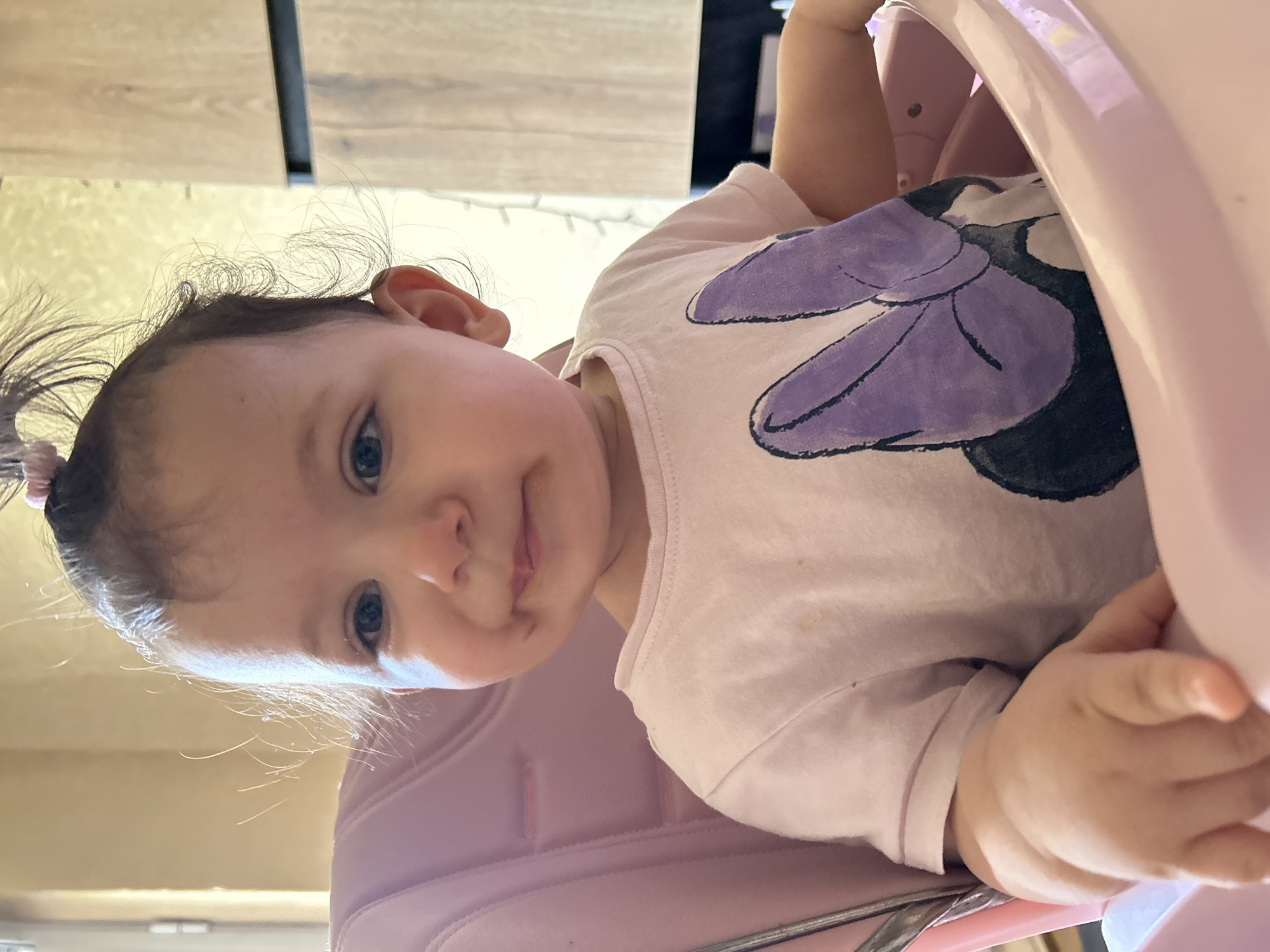 A baby with dark hair pulled into a ponytail sits in a pink high chair. The baby is wearing a light pink t-shirt with a purple Minnie Mouse bow graphic on it. There is a small amount of food around the baby's mouth. The baby's left hand is resting on the edge of the high chair. The background shows a wooden cabinet and a window with light-colored curtains. The scene appears to be an indoor domestic setting, possibly a kitchen or dining area in Sofia, Bulgaria.