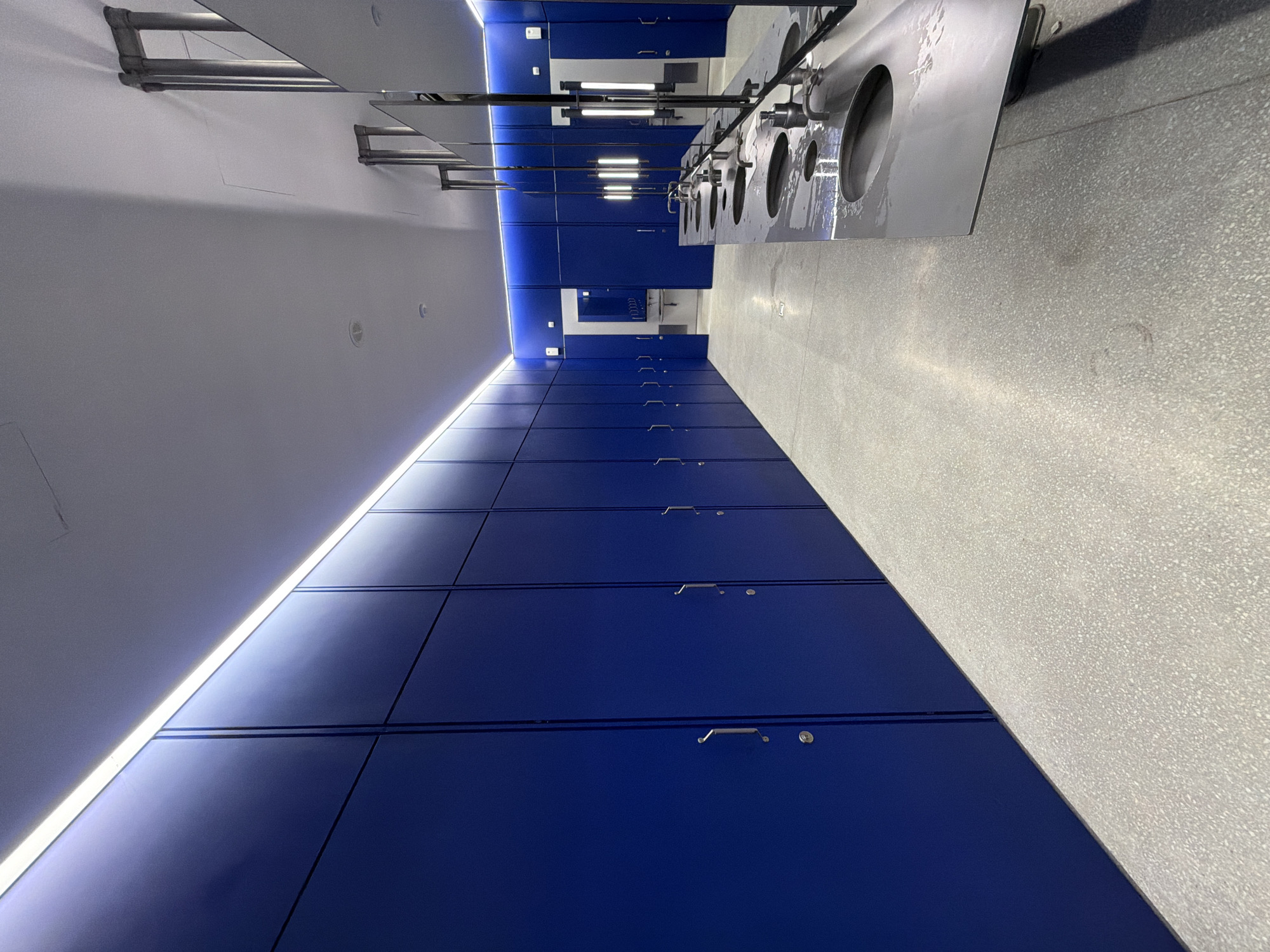 The image presents an interior space featuring a row of blue lockers running along the lower half of the frame. Above the lockers is a white ceiling with an integrated lighting strip creating a bright line along the lockers. Along the upper part of the frame are several stainless steel sinks. The floor is a speckled gray. A deeper section is visible further in the space, suggesting the end of the row of lockers. The overall perspective suggests an institutional or public facility. The image does not allow for determining its location in Moscow, Russia.