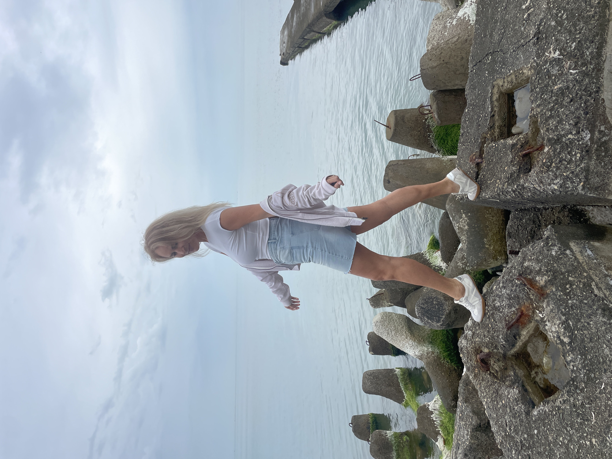 A Caucasian woman with blonde hair stands on concrete blocks at a breakwater. She wears a white shirt, light blue denim skirt, a white jacket draped over her shoulders, and white sneakers. Her arms are outstretched for balance. The concrete blocks are adjacent to the sea, and moss grows on them. The scene is located at a breakwater in Sochi, Russia.