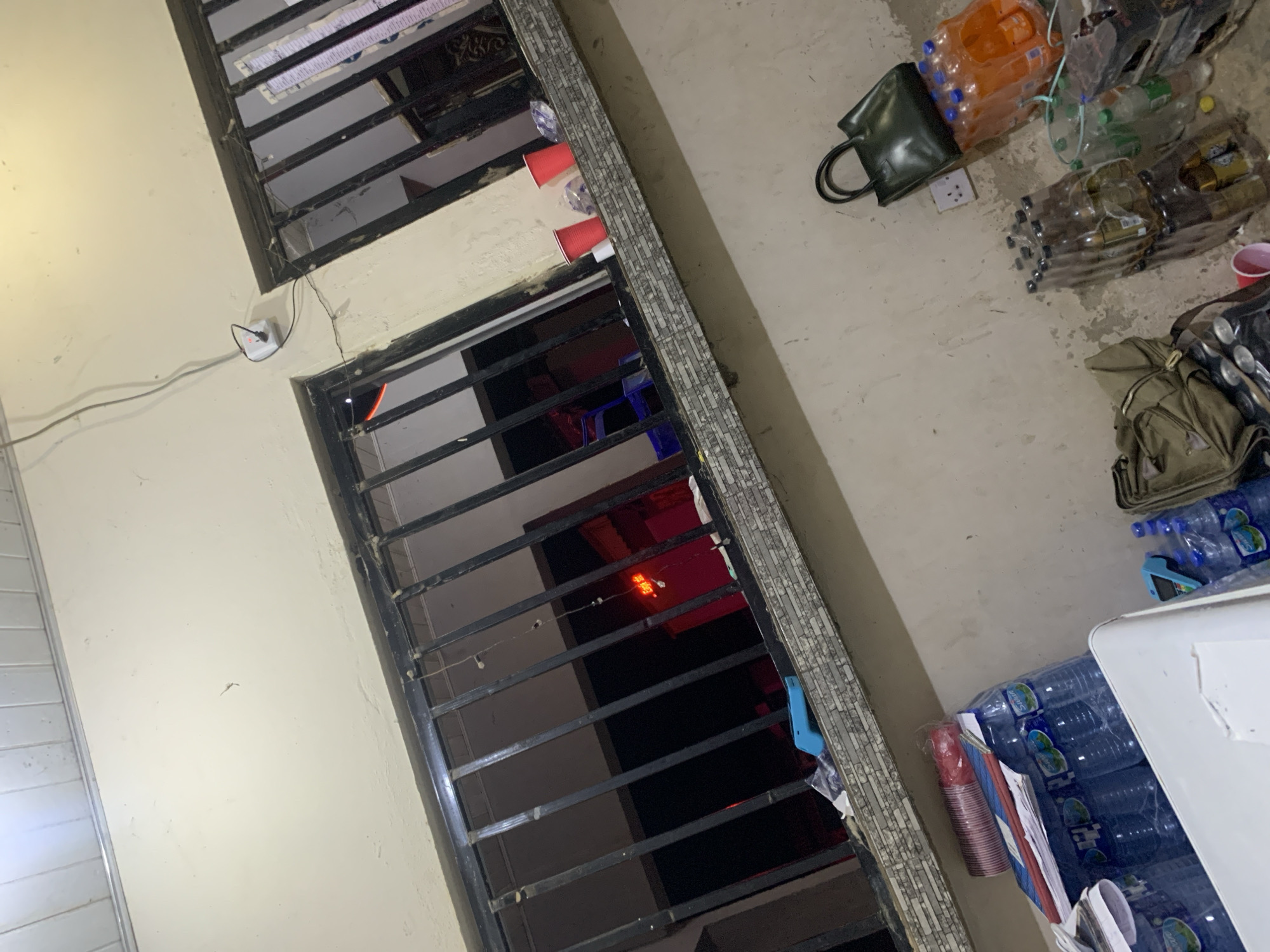 The image depicts an exterior view of a building in New Stadium, Nigeria, possibly at night. Two barred windows are visible, with the left window showing a red neon sign inside. Several plastic cups, some red and some white, are placed on the windowsill of the right window, along with some plastic bags and what appears to be a roll of white fabric. On the ground level below the windows, there are stacks of plastic water bottles and orange-colored beverages. A dark green handbag rests on top of some of the bottles. Various bags and containers are scattered on the ground, along with some discarded items. The overall impression is one of an outdoor storage or informal gathering area.