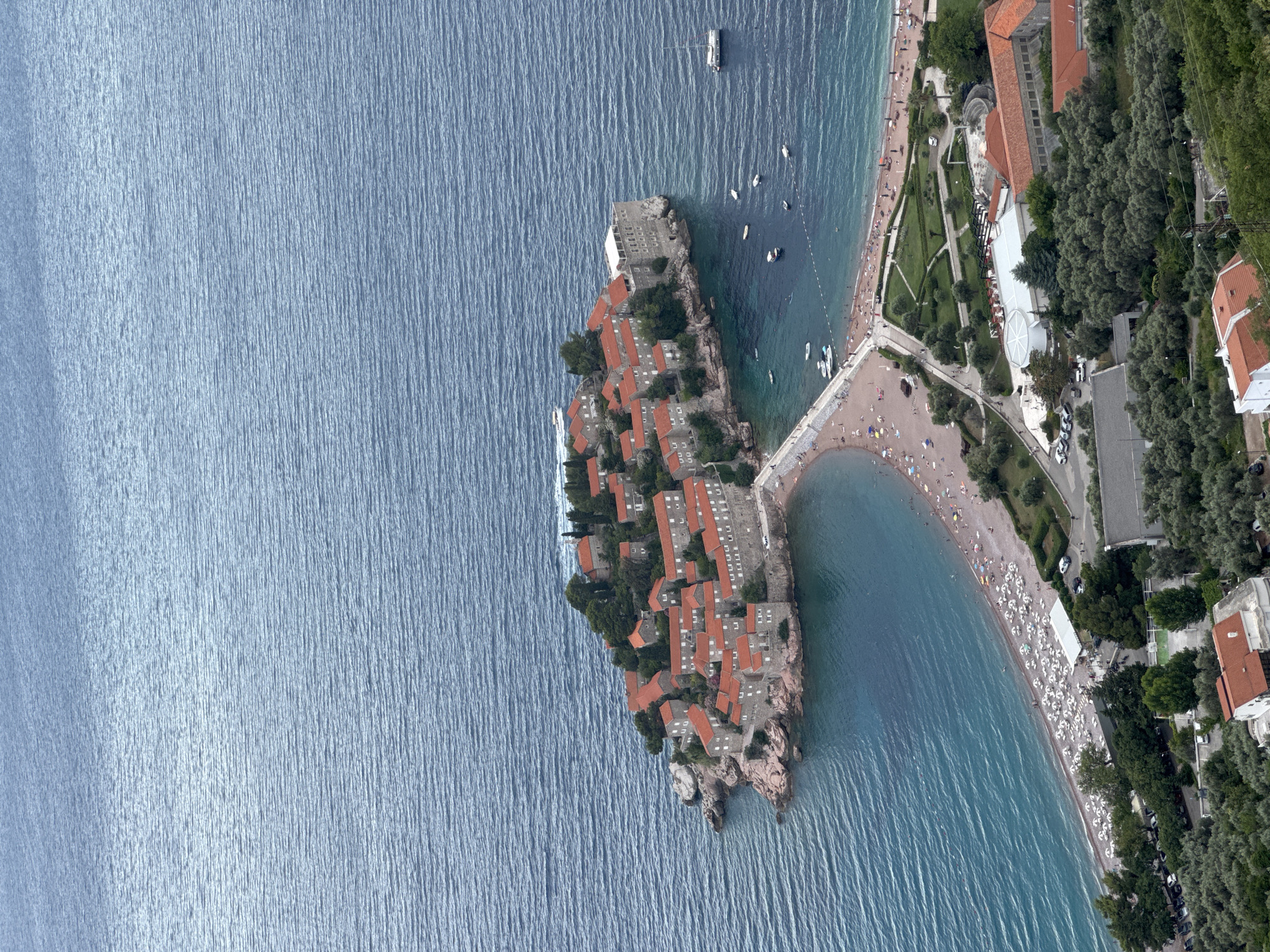 This is an aerial photograph of Sveti Stefan, Montenegro, showcasing the picturesque island resort connected to the mainland by a narrow causeway. The turquoise waters of the Adriatic Sea gently lap against a pebble beach dotted with numerous white umbrellas and lounge chairs, indicating a leisurely day at the seaside. On the beach, several people can be seen relaxing and enjoying the sun.
The island itself is a cluster of ancient stone buildings with distinctive red-tiled roofs, nestled amongst lush green trees. These buildings form a charming village-like atmosphere. The causeway, also crowded with people, serves as a vibrant pedestrian walkway. Beyond the beach, a road runs alongside the coastline, with parked cars and more greenery. The overall scene suggests a bright, clear day, ideal for a vacation. The architecture and the natural beauty of the coastline point to a Mediterranean setting, consistent with the known location of Sveti Stefan.