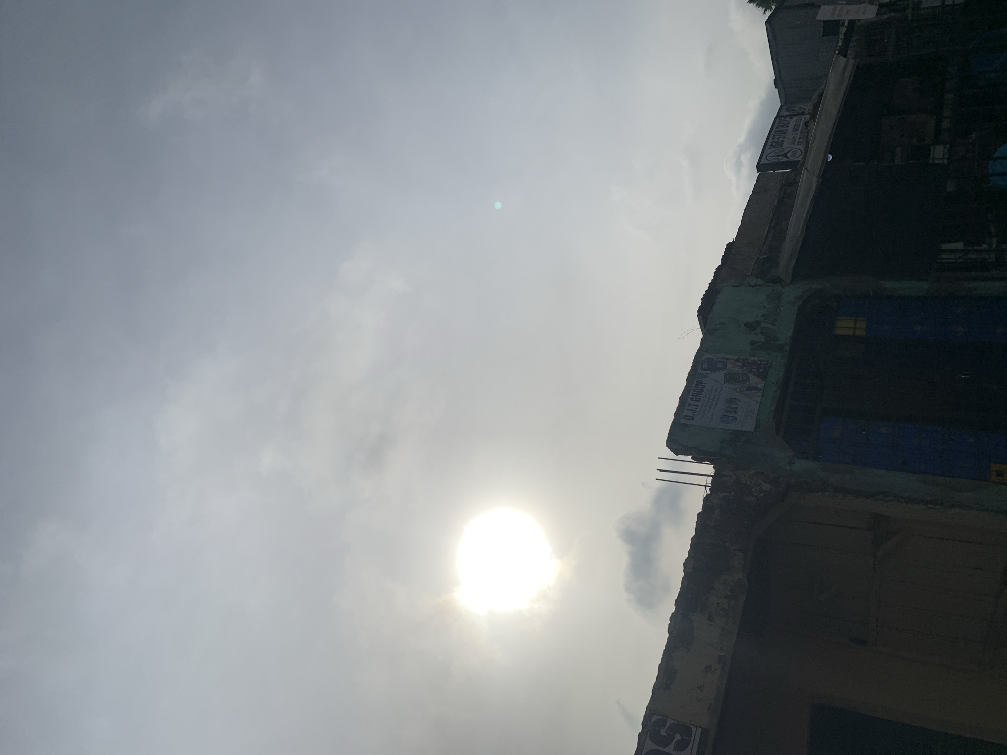 The image, rotated 90 degrees counter-clockwise, depicts a bright, cloudy daytime sky with partial views of buildings on the right. The sun is a dominant feature, appearing as an intensely bright, overexposed white orb with a strong halo, positioned towards the bottom-center of the frame. The sky is largely covered by light, diffuse clouds, and a small, faint bluish-green lens flare is visible near the upper-center.
On the right side of the frame, sections of what appear to be multi-story buildings or structures are present. These buildings exhibit a worn appearance, with visible exposed concrete, and patches of teal-green and blue paint. Reinforcing bars protrude from the top of one concrete section, suggesting ongoing construction or a dilapidated state.
Several signs and posters are affixed to the building facades. A clear white banner near the top-right displays the text "D.J.T GROUP." Below this, another sign is partially visible, showing indistinct text that could include "STAR CAFE" or "STAR HOTEL." At the very bottom edge of the frame, part of a sign showing "SO" or "SQ" is visible.
No people are present, and no specific activity is taking place. The bright sun indicates it is daytime, likely midday or early afternoon, with a cloudy but luminous sky. The architecture and visible text are consistent with an urban or semi-urban setting in Rafin Par, Nigeria.