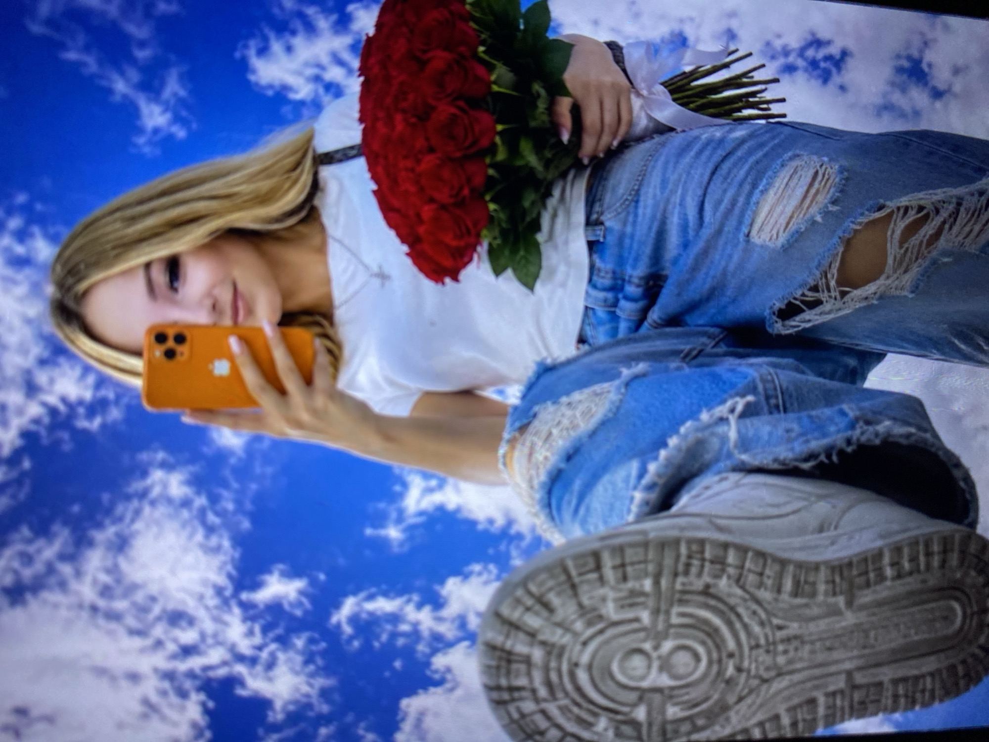 The image is an eye-level, medium shot of a young blonde woman holding an orange smartphone in her right hand, taking a selfie. She is wearing a white t-shirt, ripped blue jeans, and white sneakers. She is also holding a large bouquet of red roses in her left arm. The background is a bright blue sky with white clouds. The image was captured indoors, possibly in a studio, as indicated by the artificial sky backdrop. There are no identifiable landmarks or clues to suggest the location is the city of Luh, Ukraine.