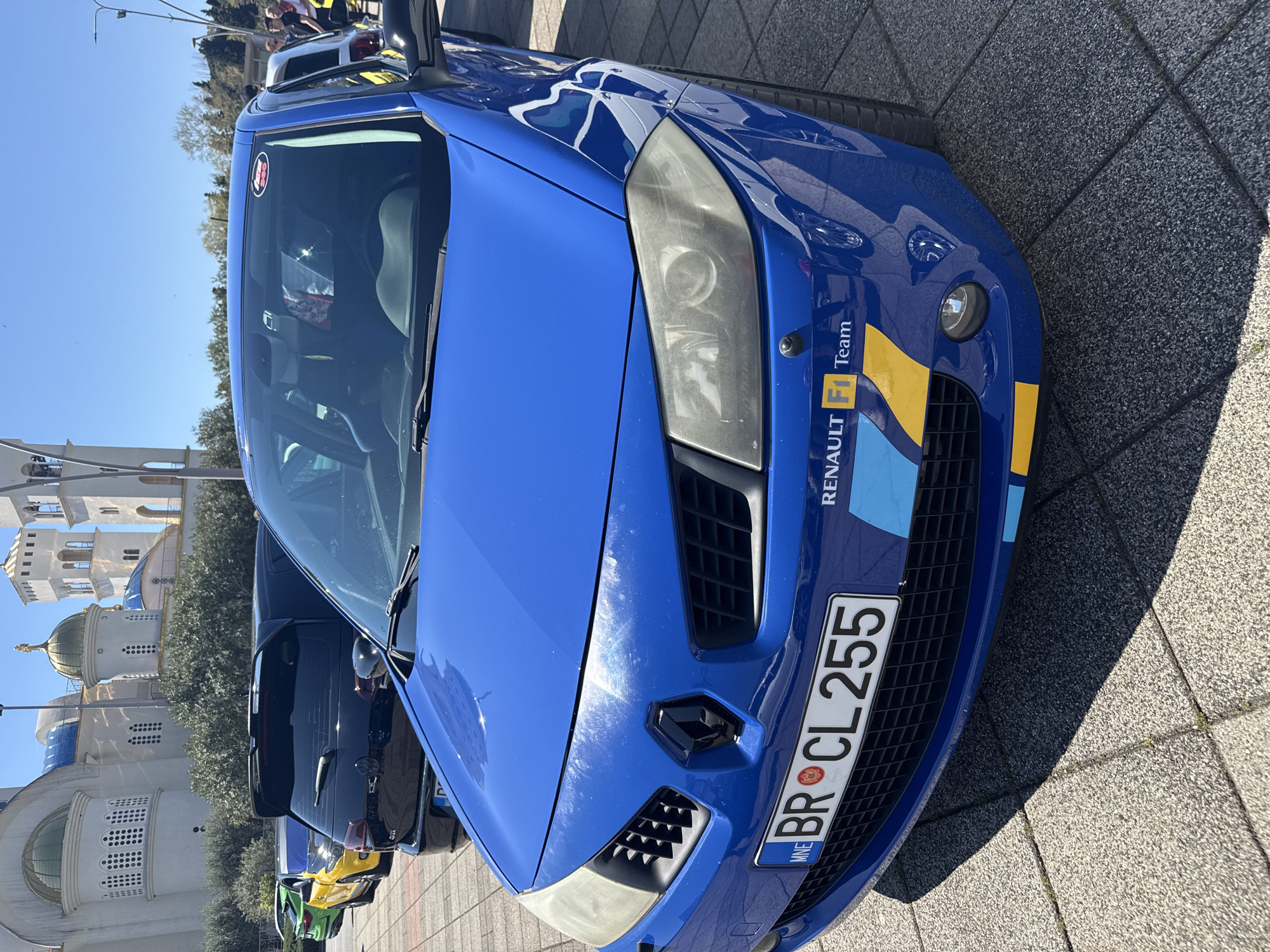 The image shows a blue Renault Megane parked on a tiled surface, likely a pedestrian area, in an urban environment. The car has "RENAULT F1 Team" branding on its front bumper and the license plate reads "BR CL 255", indicating it is registered in Montenegro (MNE). To the left of the car, there's a line of other parked vehicles in various colors (green, yellow, and black). In the background, there is a view of an Eastern Orthodox church-like building with domes and towers, set against a clear blue sky. The location is unknown, Montenegro.