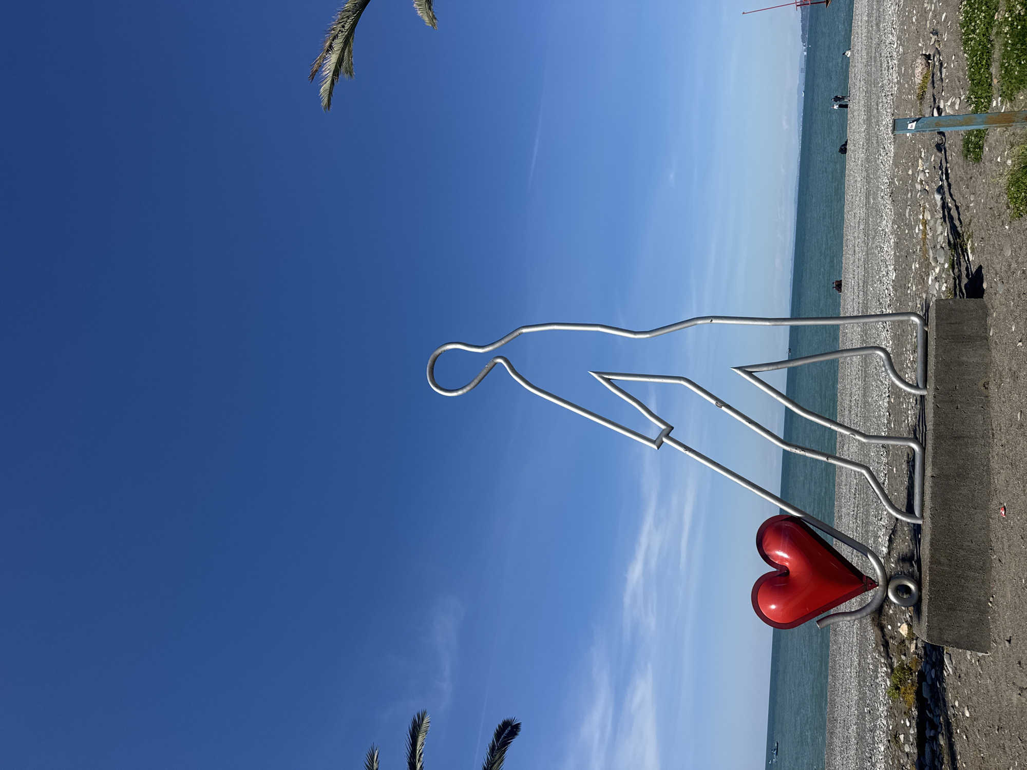 A metal statue stands on a concrete base next to a beach. The statue depicts the outline of a person pulling a red heart on a platform. The beach has small pebbles and is next to a body of water. The sky is blue with some wispy clouds. Palm leaves are visible at the top and bottom of the frame. Location: Unknown, Unknown.