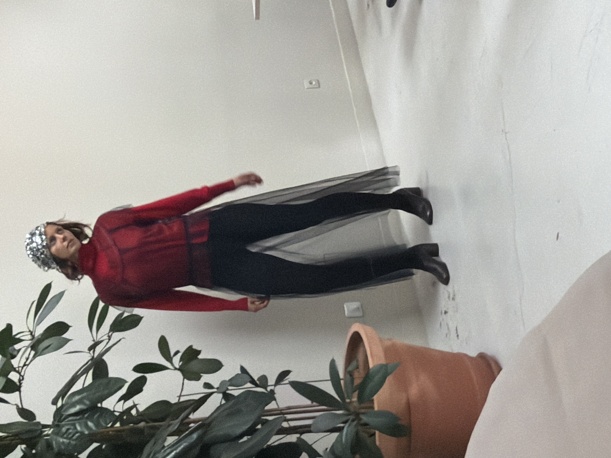 A woman is dressed in a red top, black leggings, and black boots, along with a sparkly headpiece, and is lying on the floor. She is positioned near a potted plant with dark green leaves and a large clay pot. A white wall is behind her, with a light switch visible. The setting appears to be indoors, possibly a residential space. It is not possible to determine the location in Paris from the image.
