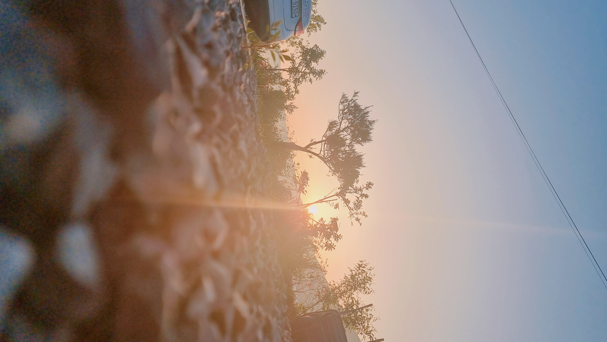 The image depicts a landscape scene with the sun low on the horizon. A silhouetted tree stands against the bright light of the sun, partially obscuring it. In the foreground, there is blurred terrain, possibly rocks or dirt. In the upper portion of the image, a car is partially visible. An overhead power line crosses the clear sky in the background. The scene appears to be in Dunnae-myeon, South Korea, and the image is oriented sideways.