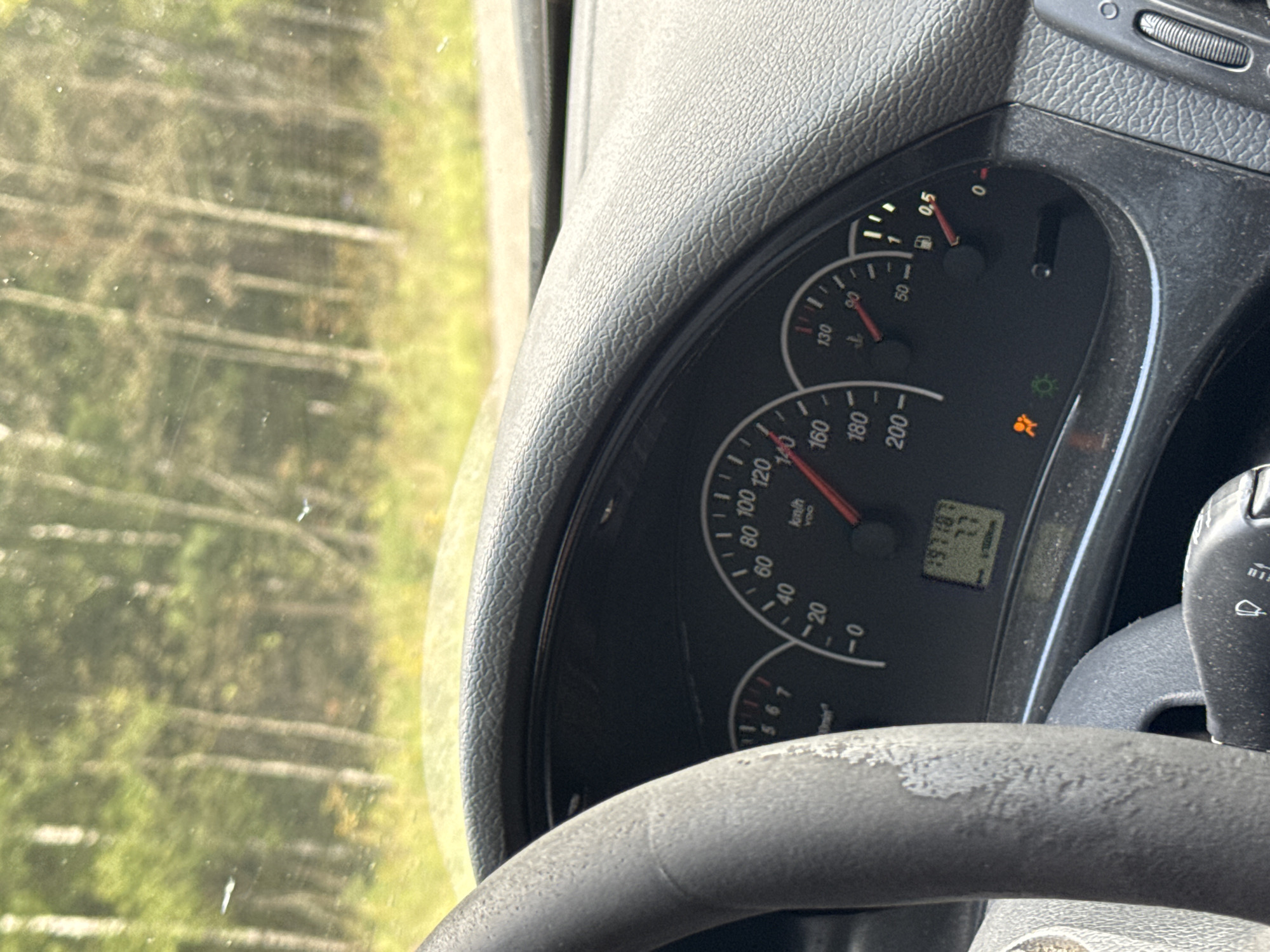 The image shows the interior of a car, specifically the dashboard and steering wheel, with a view of trees and greenery through the windshield. The dashboard features a speedometer, tachometer, fuel gauge, and an odometer displaying "197187 7.7". There are also warning lights illuminated on the dashboard, including an airbag warning light and a green indicator light. The steering wheel appears worn and dusty. The environment outside the car suggests a rural or natural setting, possibly a forest or wooded area. The location is stated to be Kazanskii Kliuch, Russia.