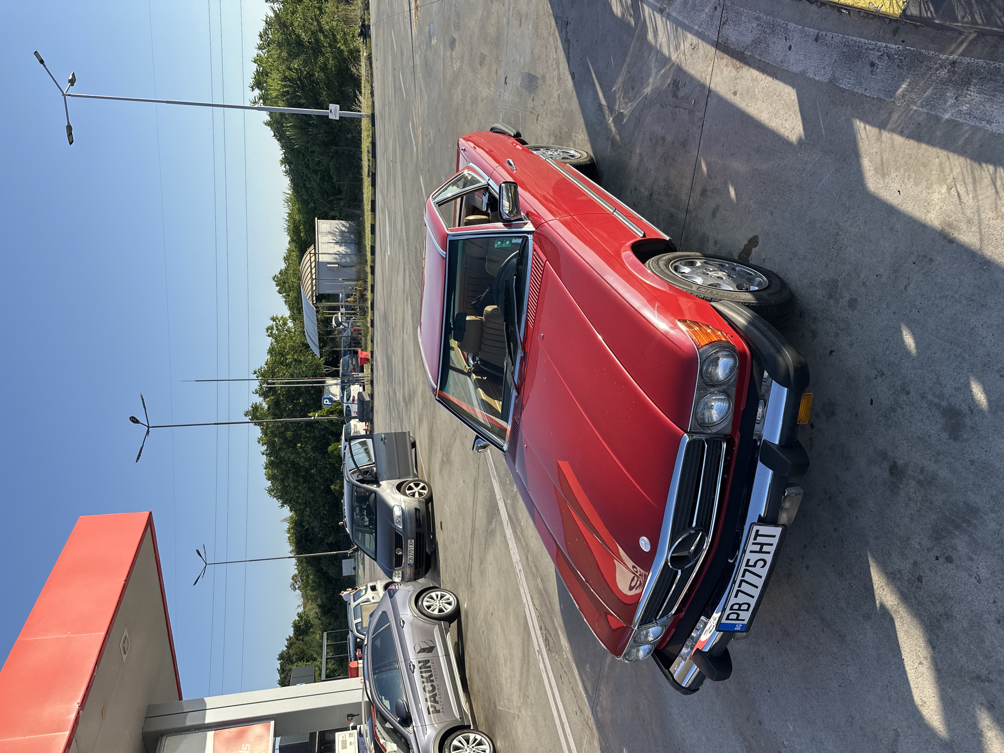The image depicts a red Mercedes-Benz SL-Class convertible parked in a paved lot, likely a gas station, under a clear blue sky. The car, with its chrome accents and distinctive Mercedes-Benz grille, is the primary focus. In the background, several other vehicles, including a grey SUV with "PACKIN PACKAGING INDUSTRY" branding and a silver Volkswagen minivan with its doors open, are visible. A red gas pump canopy with a white support column is partially visible on the left. The environment suggests an outdoor setting with trees and streetlights in the distance. The license plate of the red Mercedes-Benz reads "PB 7775 HT." The scene appears to be during daylight, with shadows cast by the car and structures indicating the sun's position.