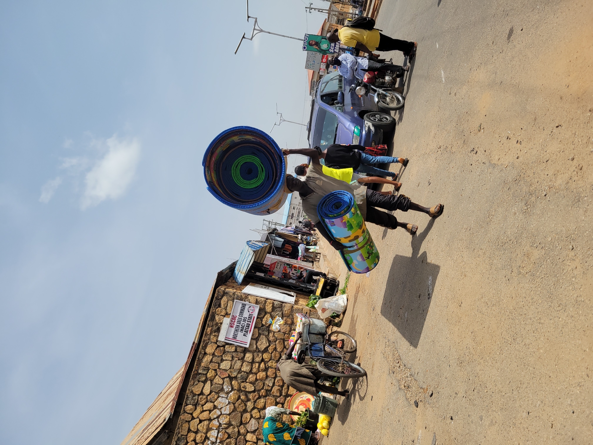 The image shows a street scene in Jos, Nigeria. A man carries a stack of rolled-up mats on his head and another rolled mat in his hand. People are standing on the sidewalk and near a blue car. A motorcycle is parked near the car. A woman sits next to a bicycle with baskets filled with goods. A stone building is on the left side of the image with a sign that reads "NSCOC (NAC) JOS PLATEAU STATE NORTHERN AREA COMMAND". Another sign reads "Opay" and "Sca Aler". The sky is blue with few clouds. The road is dusty.
