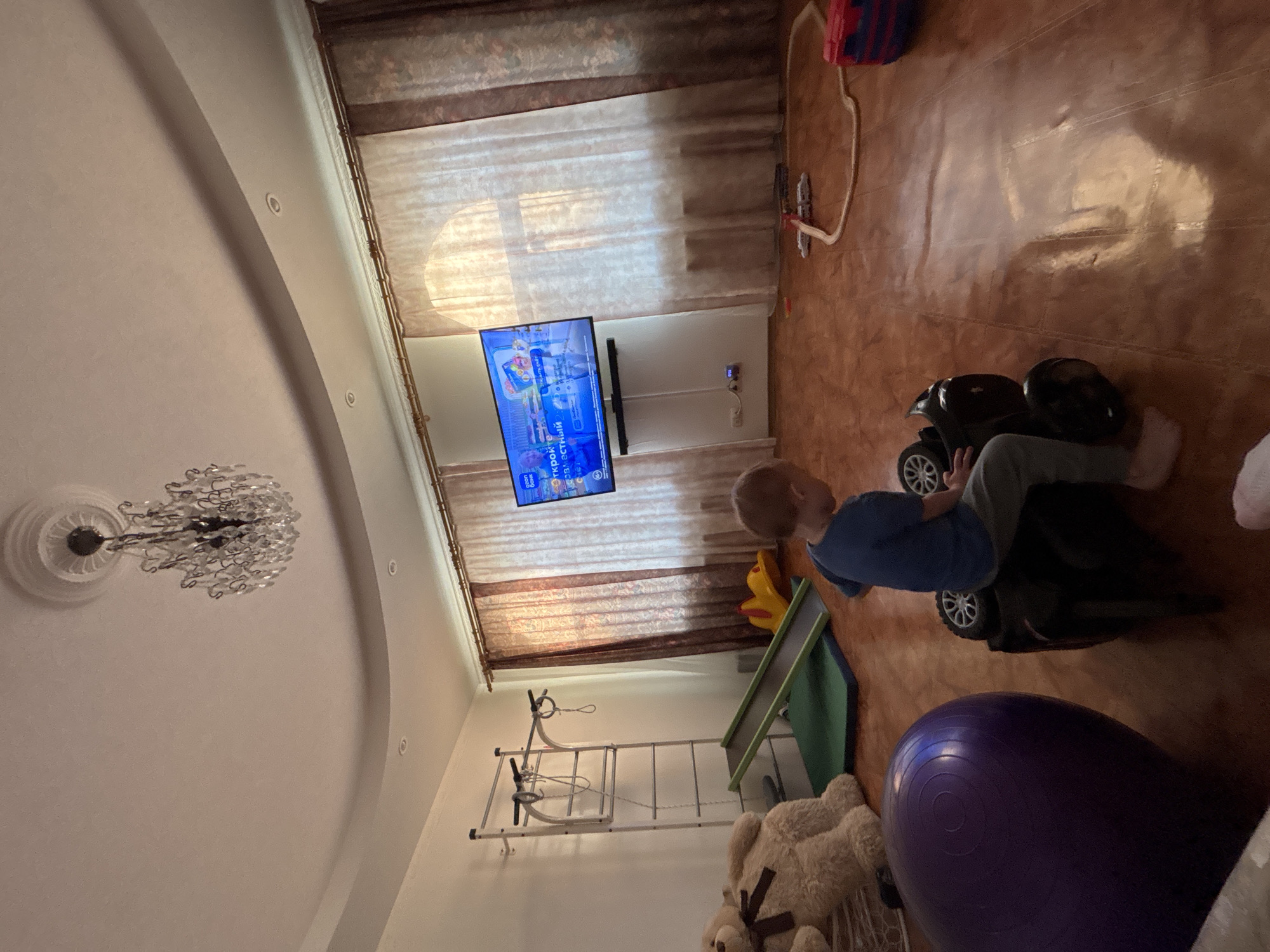 The image shows an indoor scene in what appears to be a living room, possibly in a residence in Ферма, Russia. A young boy is sitting on a black toy car, facing a wall-mounted television. The television is on and displaying a brightly colored video. Behind the boy, in the bottom right of the frame, is a large purple exercise ball and a teddy bear. Towards the wall is a children's climbing frame and slide. The room has brown tile flooring, and light colored walls with dark curtains partially covering a window. A chandelier hangs from the ceiling, which has a rounded arch design.