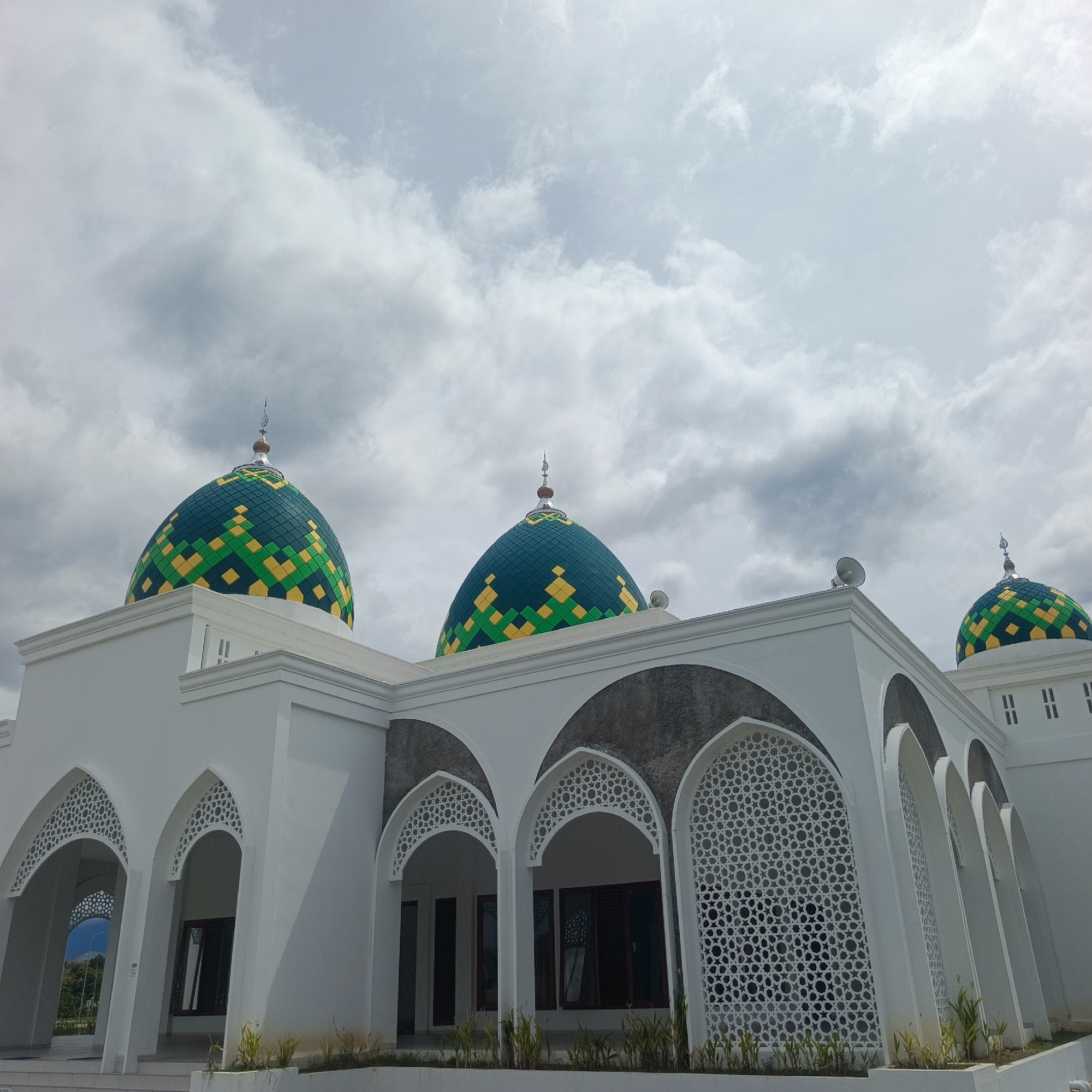 This image depicts the exterior of a mosque in Keureuweungkrueng, Indonesia. The mosque is a modern white building with several prominent green and yellow domed roofs, each adorned with a geometric pattern. The structure features multiple arched windows with intricate white latticework. In the foreground, a few small green plants are visible. The sky above is partly cloudy with grey and white clouds. There are no people or animals visible in the image.