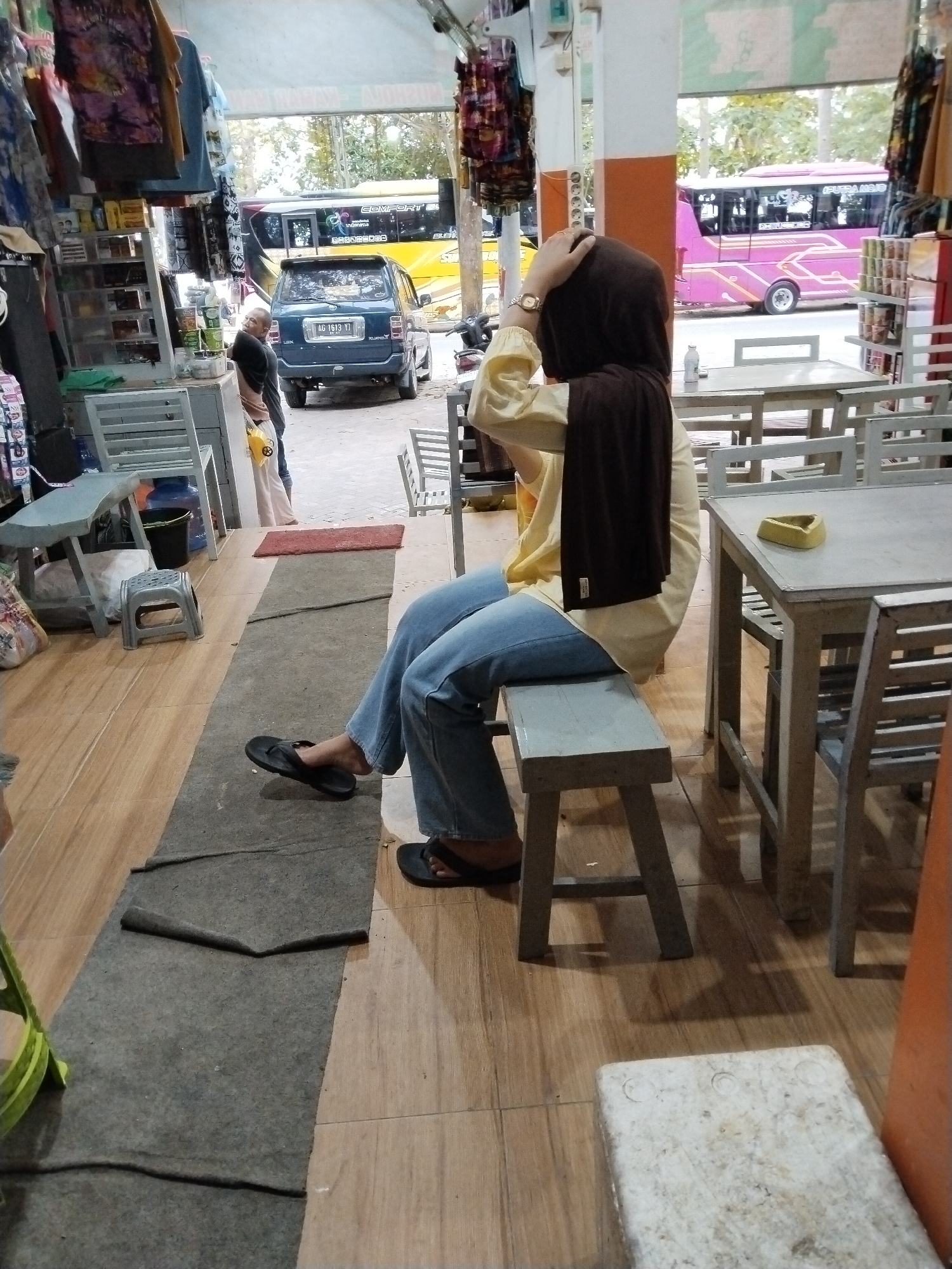The image depicts a street-side eatery or shop in Prigi, Indonesia, with an open-air setup and tables and chairs. In the foreground, a person wearing a brown hijab, a yellow long-sleeved shirt, and blue jeans is seated on a low bench, facing away from the camera and toward the street. Their right hand is touching the top of their head. The person is wearing black flip-flops.
The environment includes wooden flooring and a grey carpet runner. To the left, shelves are stocked with various items, possibly groceries or merchandise. Further down, a dark blue car with license plate "AG 1613 YD" is parked. In the background, two buses are visible on the street. One is yellow, and the other is pink with "PUTRA MAKMUR" written on its side. The scene suggests a casual, public setting, likely a stopping point for travelers.