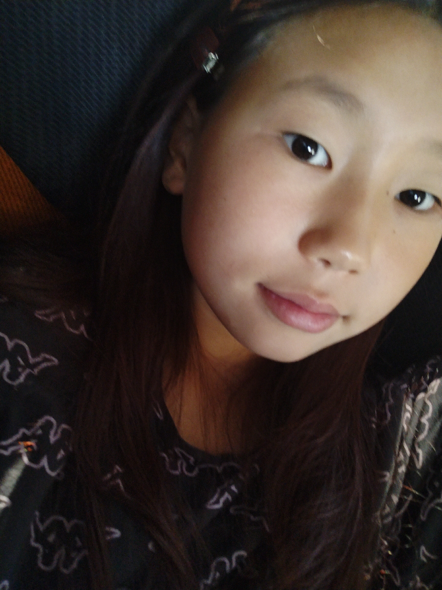 The image is a close-up, selfie-style photograph of a young East Asian female. She has long, dark brown hair that falls around her face and shoulders. She is wearing a dark t-shirt with a white, cloud-like pattern. Her skin is fair, and she has dark eyes and light pink lips. The background is dark and appears to be fabric, possibly upholstery or clothing, with a hint of orange on the left side. There is no discernible information about the location being in Turka, Russia, or any specific city.