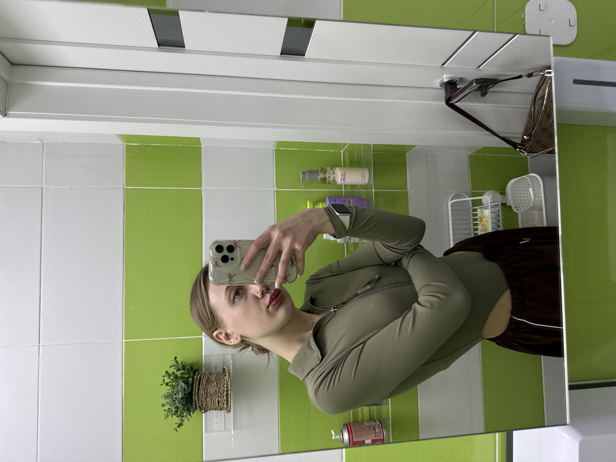A female subject is depicted taking a mirror selfie in a bathroom located in Rostov-on-Don, Russia. The subject is a young Caucasian woman with light brown hair pulled back, light blue eyes, and red lipstick. She has a philtrum piercing and a small earring in her left ear. Her fingernails are decorated with a light base and pink bow patterns. She is wearing an olive-green, long-sleeved, quarter-zip cropped top and dark brown velour sweatpants with a white stripe and logo on the left leg. A light-colored smartwatch is visible on her left wrist. She holds a smartphone with a clear case adorned with pink bows, displaying a triple-lens camera system.
The bathroom features walls tiled with alternating horizontal bands of white and bright lime green rectangular tiles. Visible in the reflection are several clear shelves holding a small woven pot with a faux plant, a pink aerosol can, and multiple pump bottles containing personal care products. To the right, a white door frame and a door with a dark horizontal accent panel are present. A brown shoulder bag with a patterned exterior and a metal chain strap hangs from the door's handle. A white wire shelving unit and a white plastic storage basket are partially visible in the background reflection. A white wall-mounted soap dispenser is visible at the bottom right of the mirror's frame.