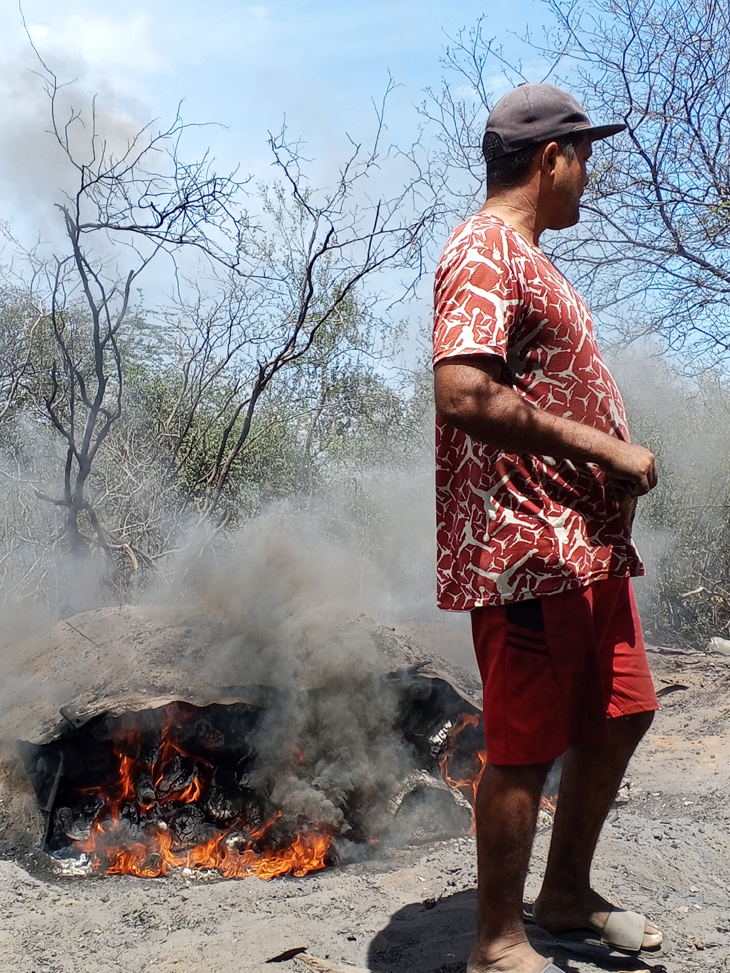 A medium eye-level shot depicts a man standing beside a fire. The fire is emanating thick grey smoke. The man is wearing a red and white patterned shirt, red shorts, and a dark baseball cap. He is also wearing light-colored slides. The environment appears to be dry and arid, with bare trees visible in the background. The location appears to be the city of Unknown, Unknown.