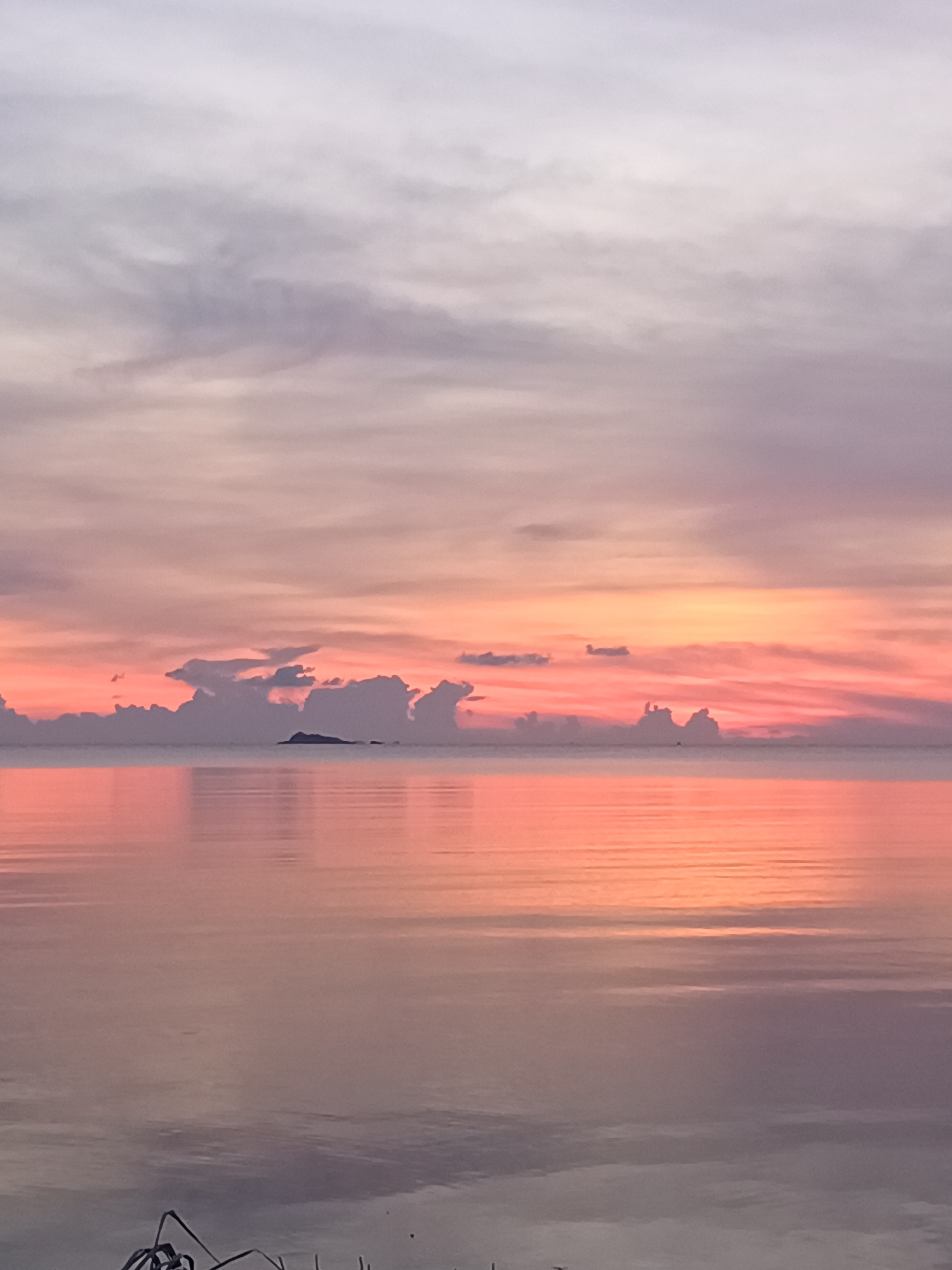 The image depicts a serene sunset over water, likely a sea or ocean, in Baan Wok Tum, Thailand. The sky displays hues of pink, orange, and gray, with clouds scattered along the horizon and in the upper portion of the image. A small island is visible in the distance. The water reflects the colors of the sky, creating a mirrored effect. Some foliage is faintly visible in the foreground, suggesting the image was taken from the shore.