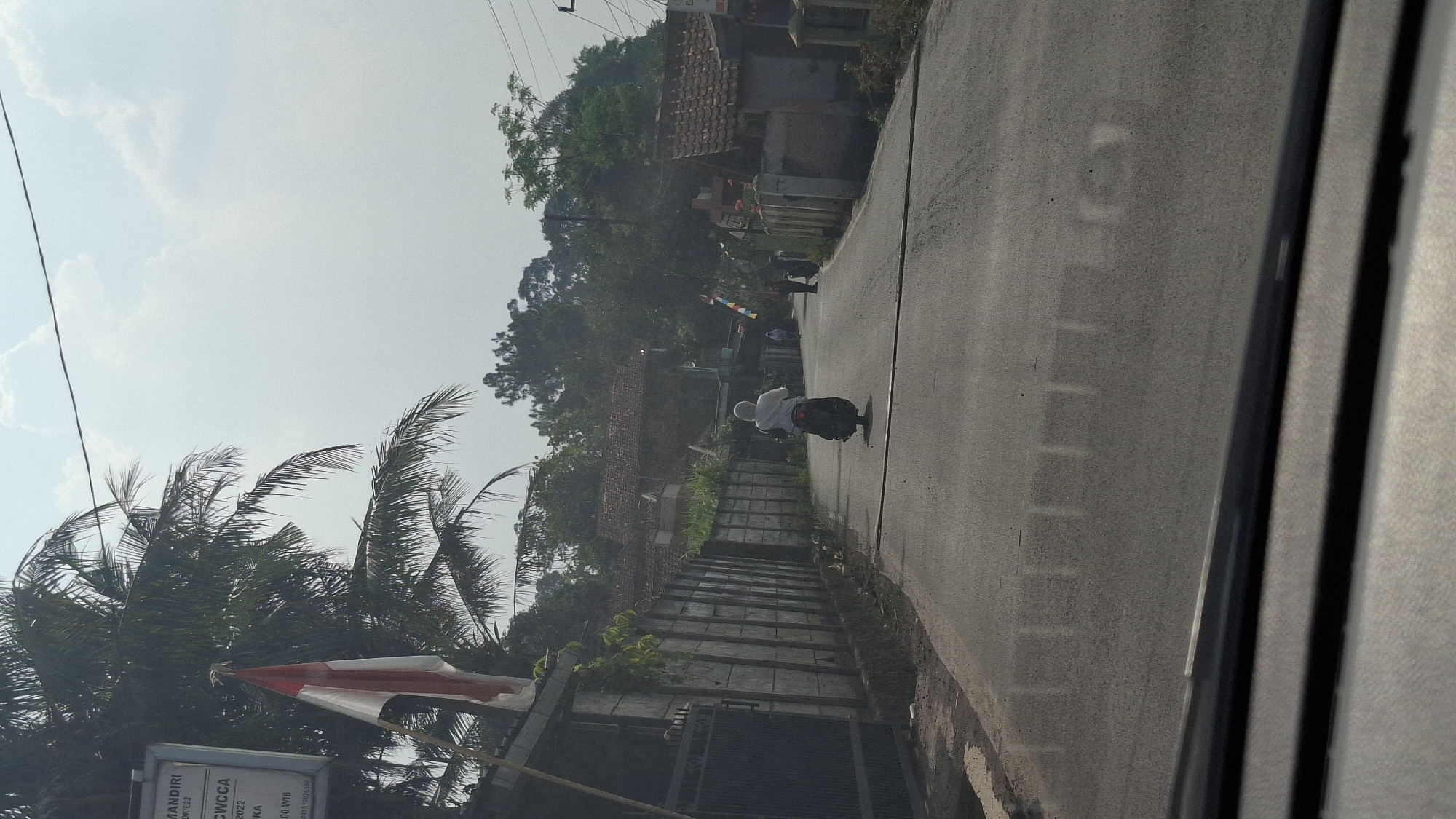 A motorcycle rider, wearing a white headscarf and long-sleeved shirt, travels down a paved road in a semi-rural area. The road is flanked by a concrete wall and palm trees on the left, and houses and trees on the right. The sky is bright and overcast. In the distance, other people can be seen walking along the road. The scene appears to be a daytime shot, possibly in Sirnagalih, Indonesia, given the presence of Indonesian flags on poles along the right side of the road. The image is taken from a low angle, possibly from inside a vehicle, as indicated by the reflection on the lower portion of the image.