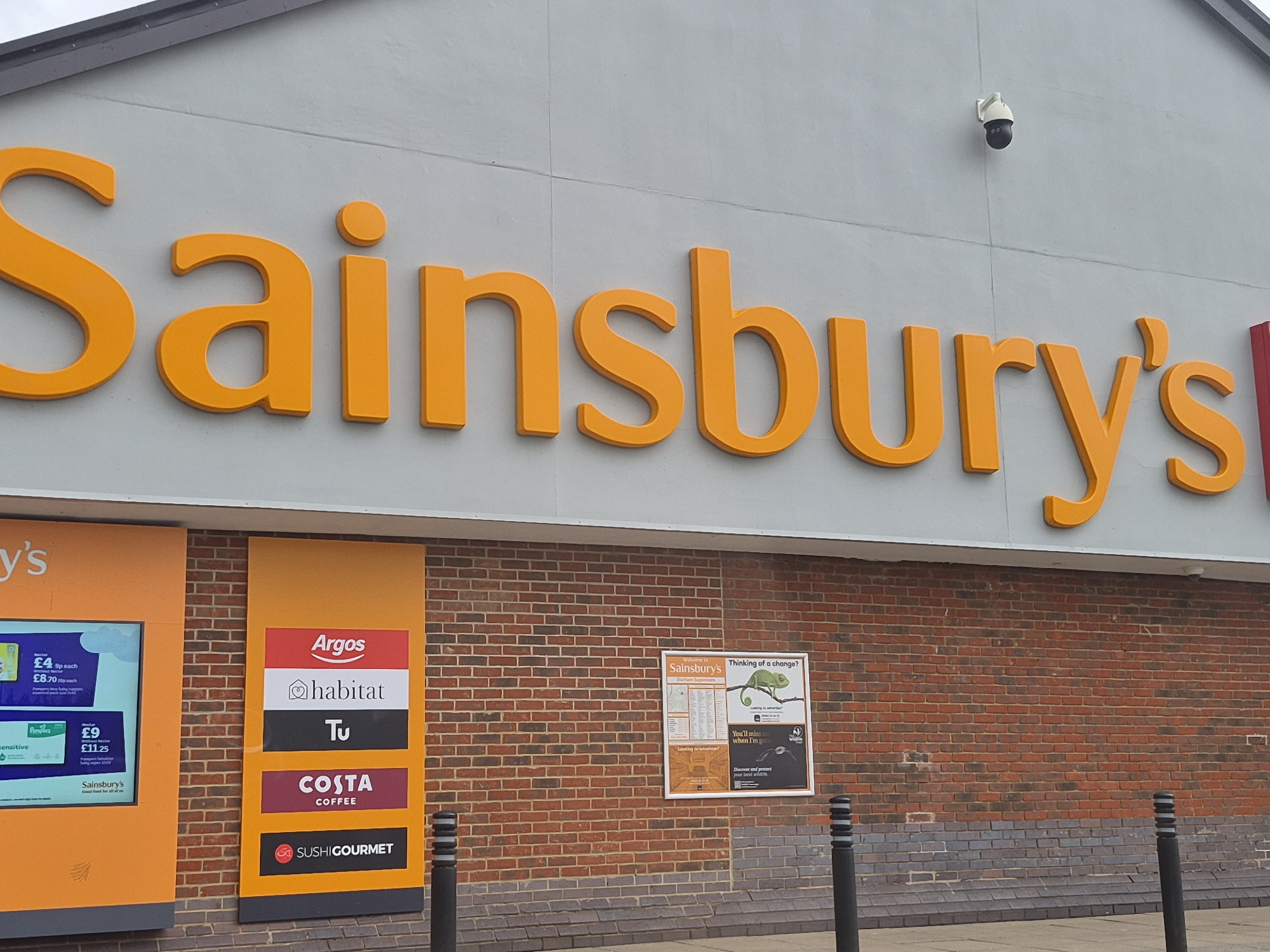 The image shows the exterior of a Sainsbury's supermarket. The large, orange "Sainsbury's" logo is prominently displayed above the entrance on a gray facade. Below the logo, the exterior brick wall features several signs. A brightly colored sign lists various services including "Argos", "Habitat", "Tu", "Costa Coffee", and "Sushi Gourmet." Another sign advertises Sainsbury's branded products, and a third poster displays a chameleon and information. A security camera is visible on the facade above the logo. Black bollards stand in front of the brick wall. The location is in Pity Me, United Kingdom.