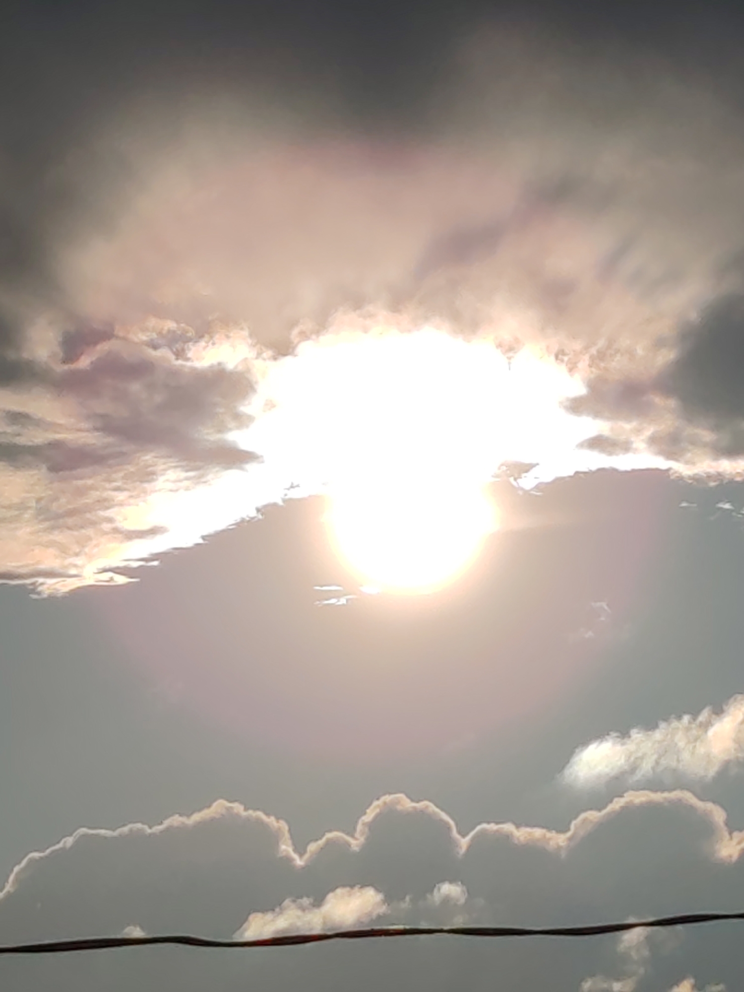 The image captures a view of the sky over Чухлэм, Russia. The sun is prominently visible, appearing as a bright, white orb partially obscured by clouds. The clouds are a mix of dark gray and white, with some edges illuminated by the sun, creating a hazy and ethereal effect. A thin, dark horizontal line, likely a power line or wire, stretches across the bottom portion of the frame. The overall lighting suggests either sunrise or sunset, with a diffused and warm glow emanating from the sun. There are no people or animals visible in the scene.