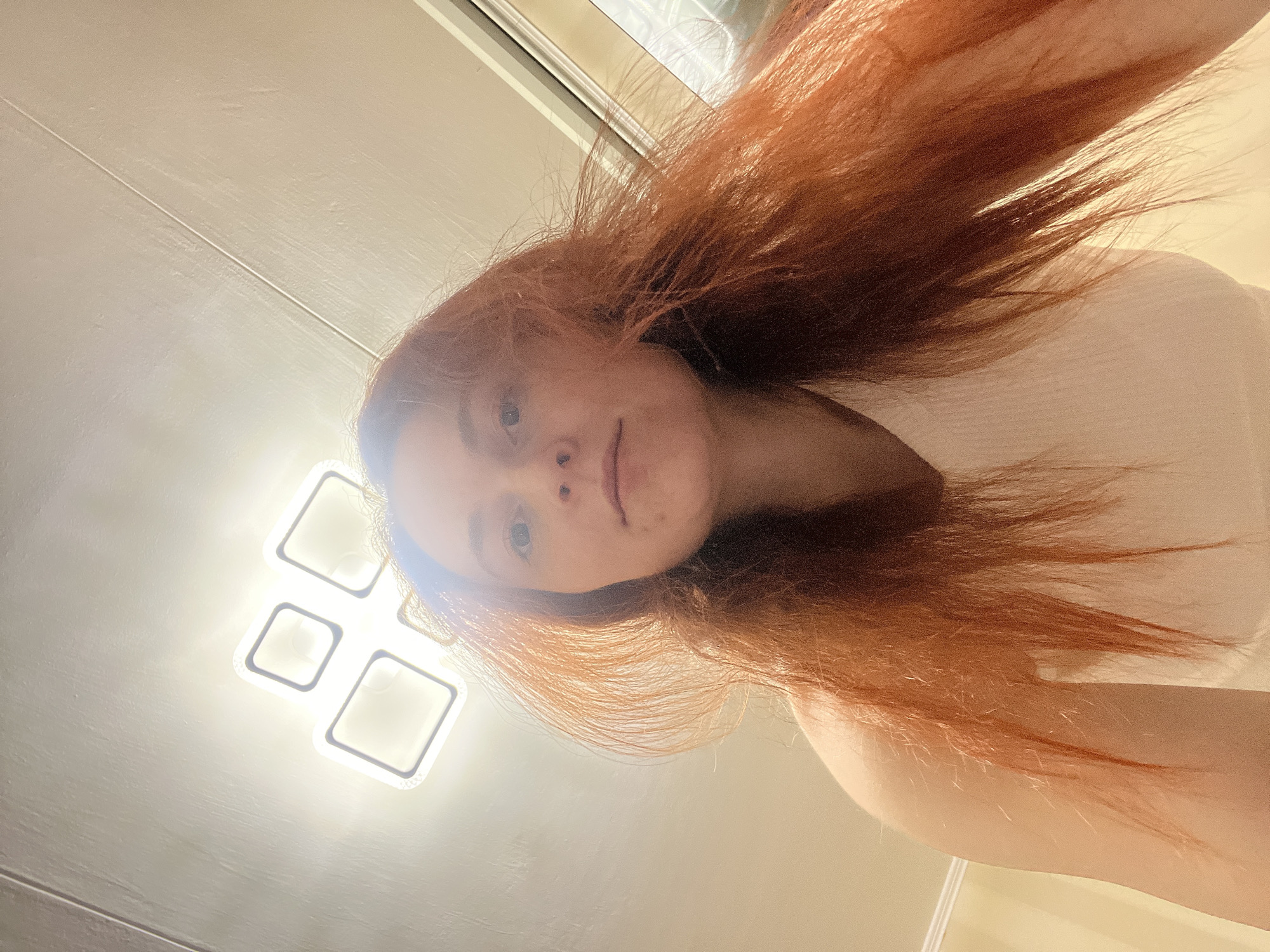 A woman with red hair is viewed from below, appearing to look directly at the viewer. The lighting is bright, with a four-panel square light fixture visible in the upper left of the frame. The woman is wearing a white top. The location is indiscernible. The city of Chaikovsky, Russia, is not identifiable within the scene.