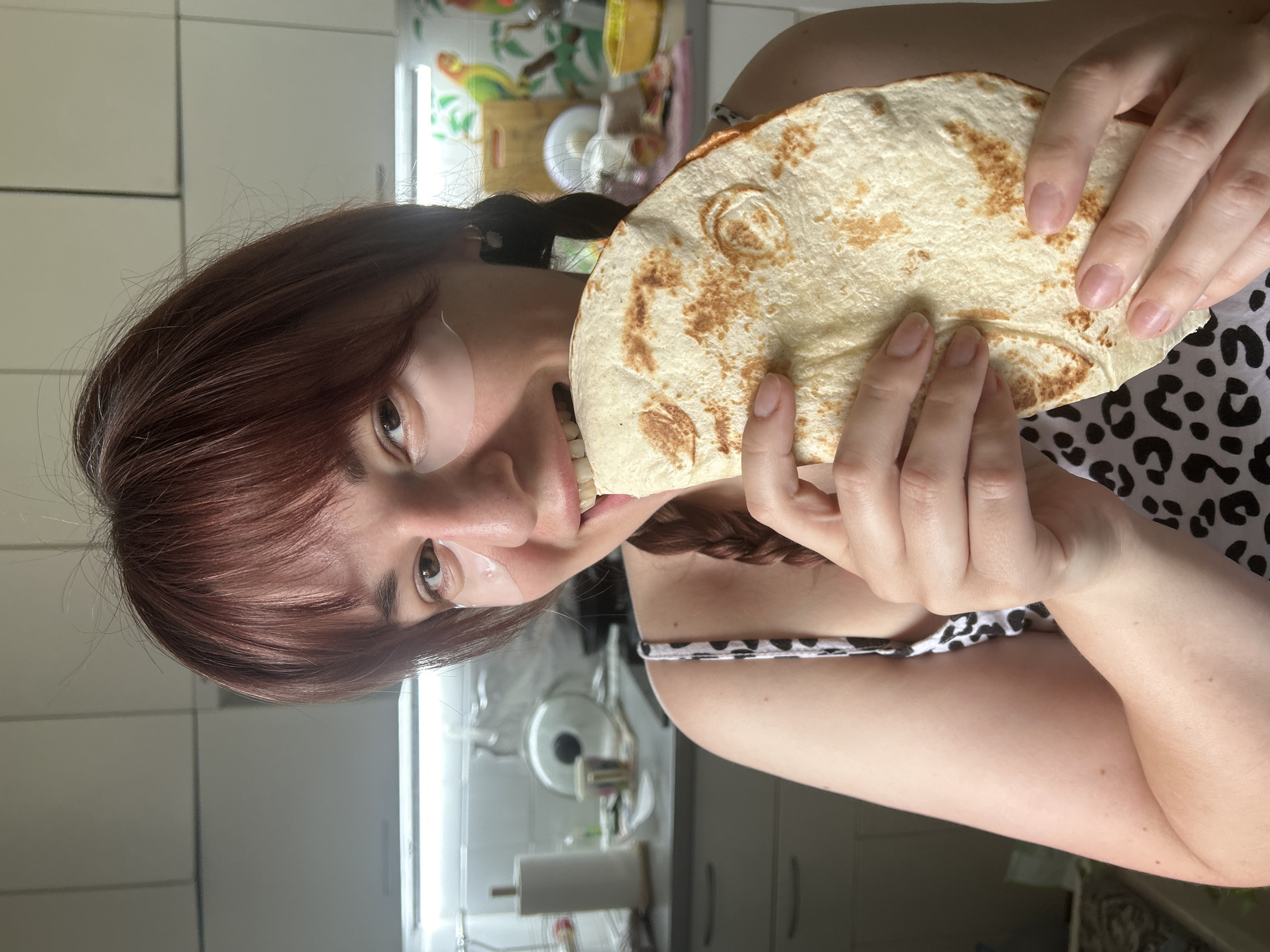 A woman with auburn hair and a black and white spotted tank top is holding and eating a flatbread in what appears to be a kitchen. She has eye patches on her face. The kitchen is brightly lit with white cabinets and appliances visible in the background. There is no indication that this image was taken in the city of Фоки, Russia.