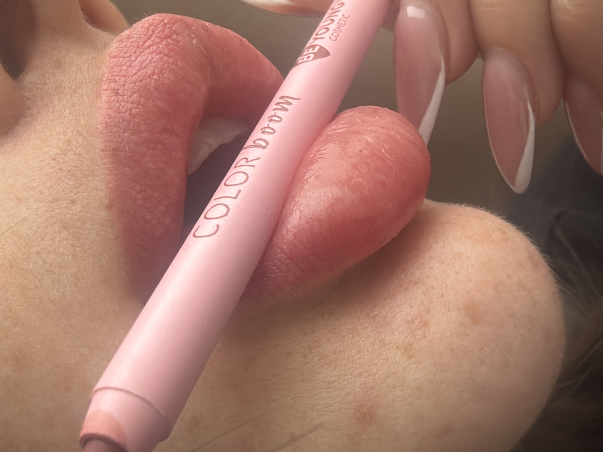 A close-up photograph depicts the lower facial region of an individual, primarily focusing on the mouth, chin, and a hand. The subject's skin is fair-toned with visible pores and scattered freckles. Fine, light facial hair is present on the skin surrounding the lips. The lips are full, exhibiting a reddish-pink hue, and are slightly parted, revealing a dark oral cavity. Natural lines and minor irregularities are visible on the lip surface. A light pink cosmetic pencil, approximately 1 cm in diameter, is held horizontally across the mouth, resting on the lower lip and positioned just below the upper lip. The pencil's exposed tip reveals a lighter pink product. Inscribed on the pencil in brown text are the words "COLOR boom," followed by "BE YOUNG" and "COSMETIC" in smaller lettering. A hand with manicured, almond-shaped nails, featuring a white French tip design, is visible in the lower right, gripping the pencil. The background is uniformly blurred, appearing as a soft, light brown or beige surface, offering no discernible environmental details. The scene is illuminated by soft, diffused lighting. The specific location within Saint Petersburg, Russia, or any geographic location, cannot be determined from the visual content of this image.