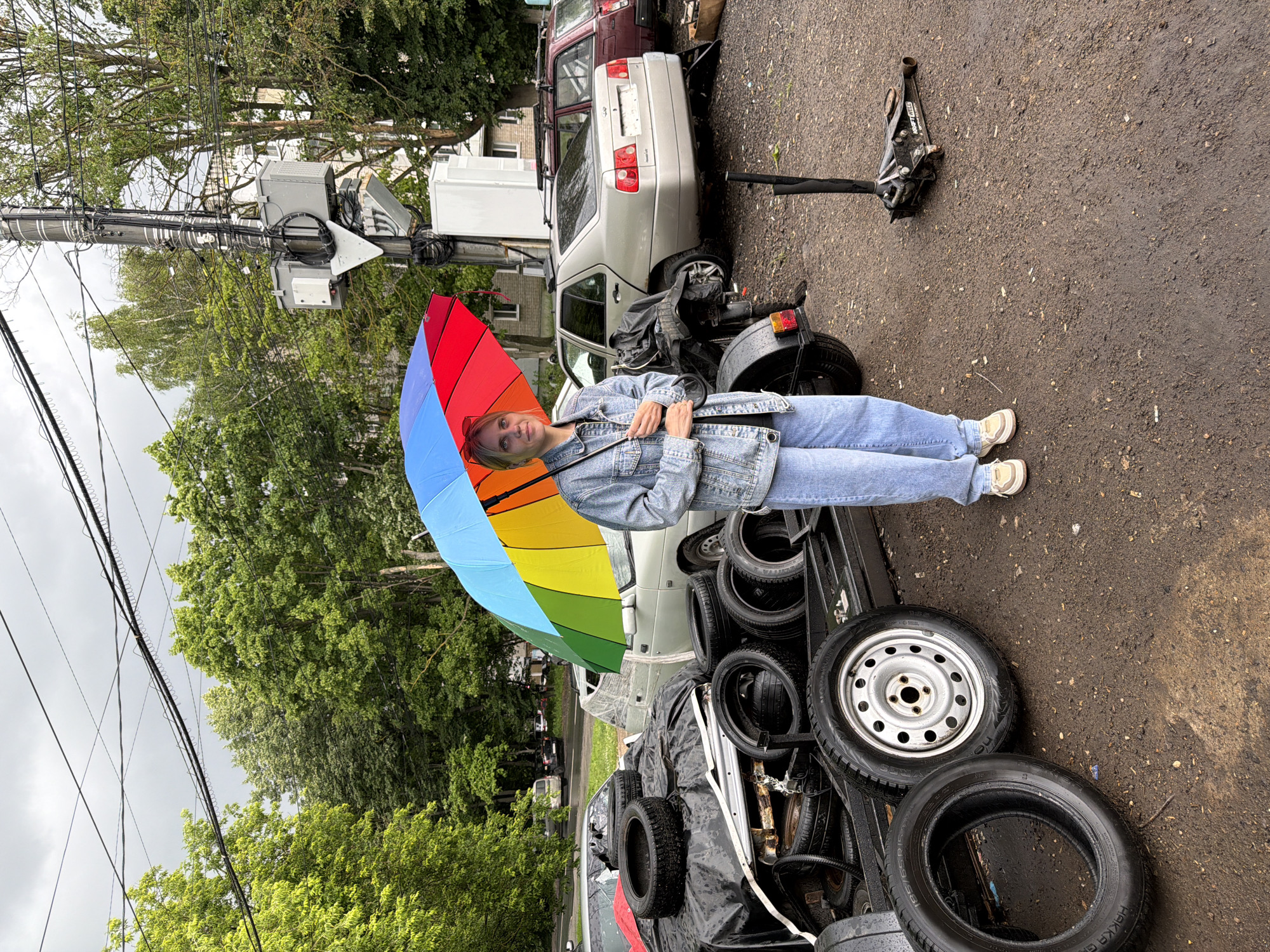 A person stands outside holding a closed, rainbow-colored umbrella. They are wearing a light blue denim jacket and light blue jeans. To their right, there is a small, dark-colored motorcycle. Behind the motorcycle and person, a light-colored sedan is parked, followed by a dark red car. The ground appears wet and muddy, suggesting recent rain or damp conditions. A stack of tires is visible in the foreground, partially covered by a black tarp. In the background, lush green trees line the scene, and a tall utility pole with numerous wires and some electronic equipment is prominent. The overall setting suggests an outdoor, possibly urban or suburban area in Nizhny Novgorod, Russia, with a cloudy or overcast sky, indicating it might be daytime, but the weather is damp. The person appears to be looking towards the camera with a neutral expression. There is no visible text in the image.