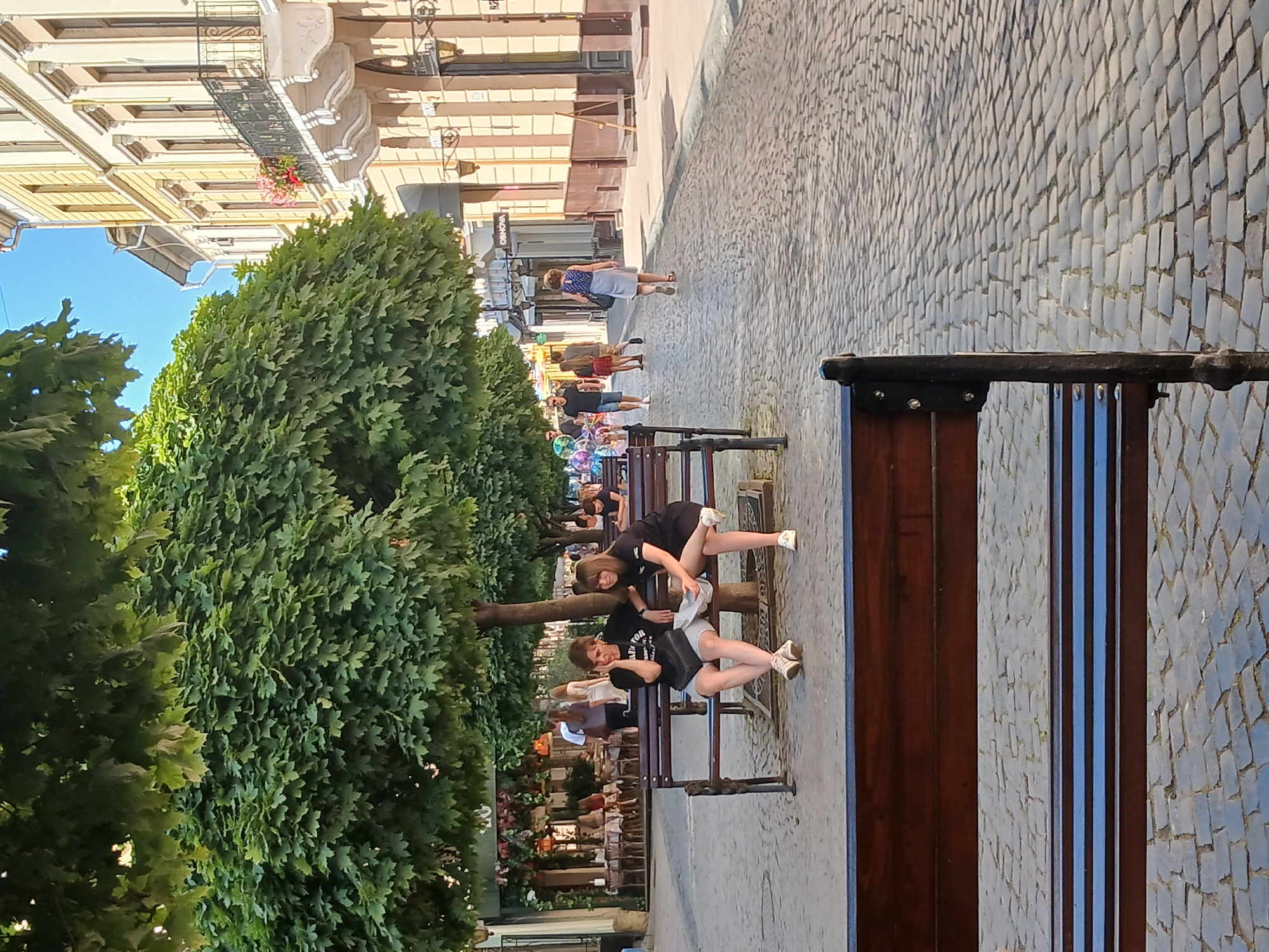 The image depicts a sunny day on a cobblestone street in an urban setting. Two young women are seated on a wooden bench in the foreground, beneath the shade of lush green trees. The woman on the left is wearing a black t-shirt, shorts, and sneakers, with her arm resting on the back of the bench and her other hand touching her face. The woman on the right is also in a black t-shirt and shorts, with her legs crossed and a white paper in her hand. Both women are looking towards the left of the frame. In the background, other people are walking along the street, and buildings with balconies and decorative architectural details line the right side of the road. Outdoor seating for a restaurant or cafe is visible on the left side of the street, under more trees. The overall scene suggests a pleasant, leisurely atmosphere.
