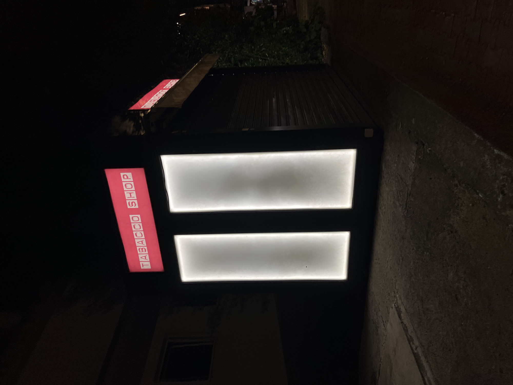 An outdoor, medium shot of a closed Tabacco Shop at night in Budva, Montenegro. The shop is illuminated by two vertical, rectangular white lights on its facade. A red sign above reads "TABACCO SHOP". The shop has a dark metal rolling shutter in front of its entrance, and is partially obscured by dense green foliage to the right. The pavement in front of the shop is worn and cracked.
