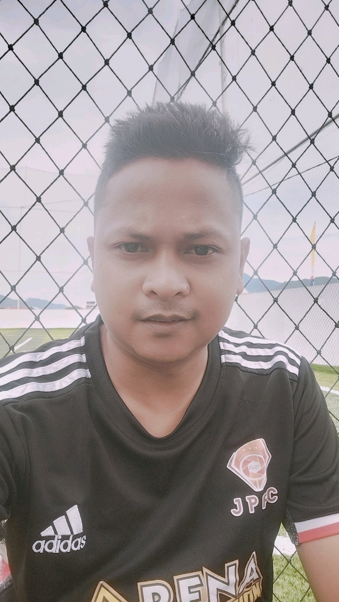 A portrait photograph shows a person in a black Adidas soccer jersey with white stripes on the shoulders. The person has short, spiky dark hair and is wearing a small ring in their nose. They are standing in front of a black mesh fence. In the background, there is a green field that appears to be a soccer field, a body of water, and a distant hilly landscape. The sky is overcast. The overall scene suggests an outdoor sporting or recreational area. The location is identified as Kalalengkio, Indonesia.
