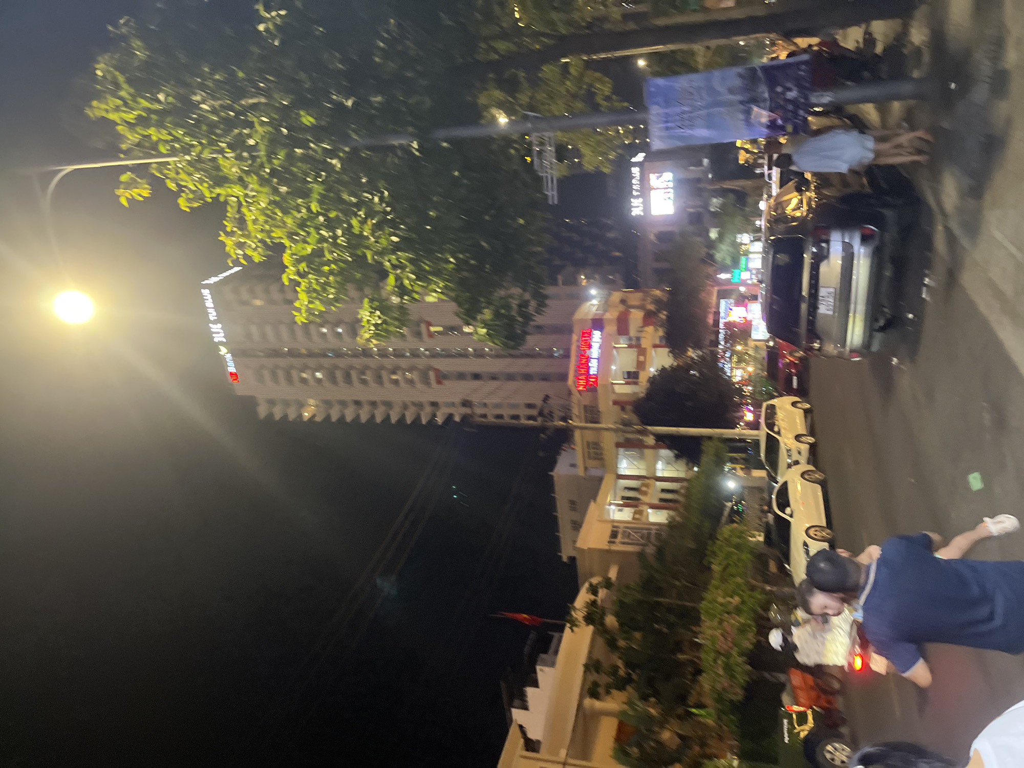 The image shows a nighttime street scene in Nha Trang, Vietnam, possibly on a main road with several buildings and trees. In the background, a tall, illuminated skyscraper with the text "BIDV BLUE TUI BLUE" is visible. Below it, a building with red and yellow signage that reads "NHÀ HÀNG - HOTEL 20 THÀNH PHỐ BIỂN" can be seen. To the right, a large vertical banner with images of the sea and possibly a resort is displayed. The street is populated with vehicles, including several cars (a white sedan, a white SUV, and a silver SUV with a license plate that appears to be Vietnamese), a motorcycle with two people on it, and another motorcycle in the foreground.
In the foreground, a man wearing a dark blue t-shirt is carrying a young child. The child is wearing a light blue garment and white shoes. Another person, wearing a white top, is partially visible on the left side of the image. A woman in a light blue top and white shorts is standing near the silver SUV on the right. The street is asphalt, and there are sidewalks with trees and streetlights. The lighting in the image is a mix of streetlights, building lights, and vehicle headlights, creating a vibrant but somewhat blurry night atmosphere.