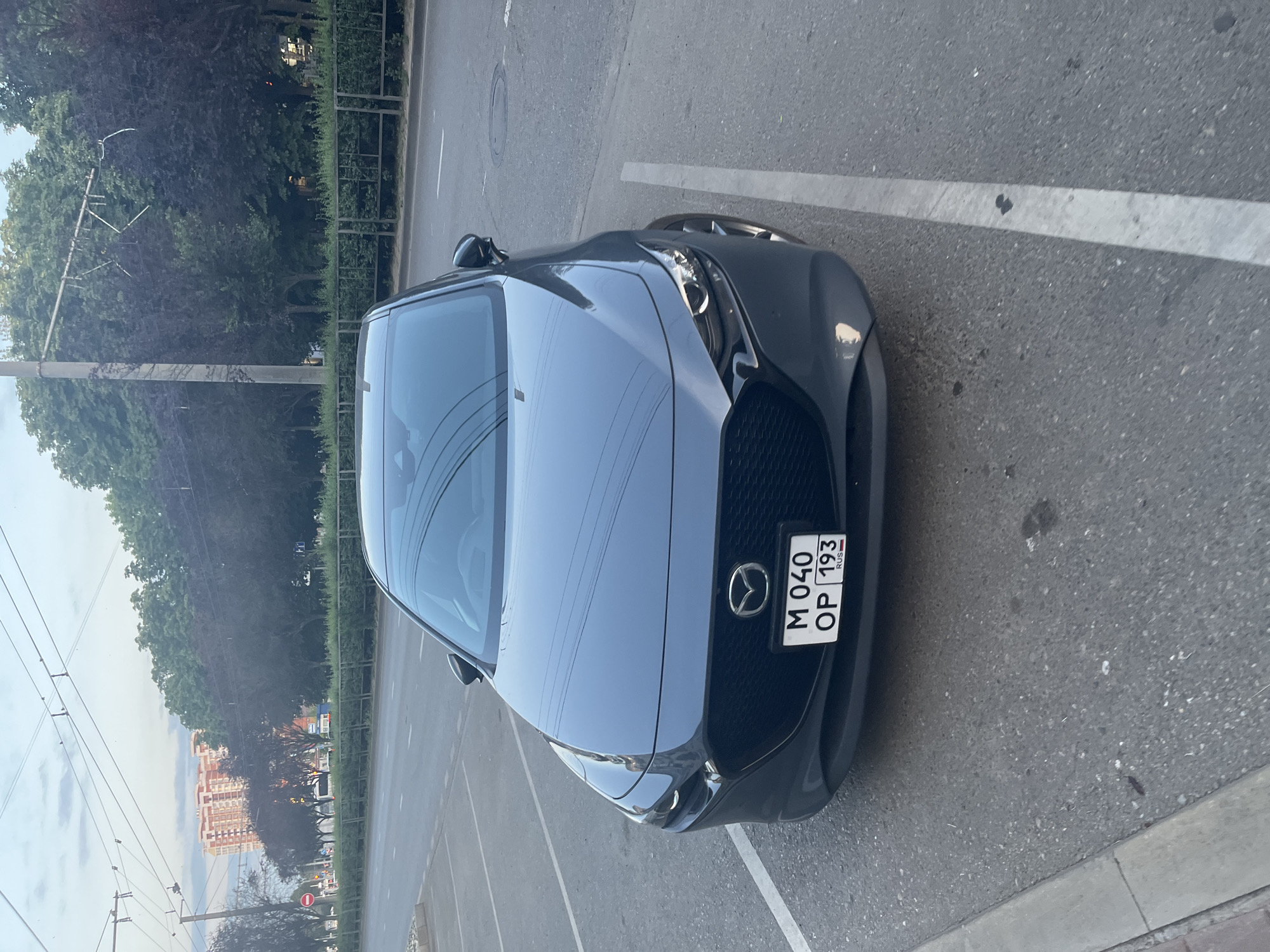 A dark grey Mazda 3 is parked in a designated parking spot on an asphalt surface. The car's front is facing the camera, and its license plate reads "M 040 OP 193 RUS". The background shows a street with trees lining the side, a sidewalk, and a multi-story building in the distance. Overhead, there are visible electrical wires, suggesting an urban environment. The sky is overcast with some visible clouds. The lighting indicates it is daytime, likely late afternoon or early evening. The scene appears to be on one of the main streets of Krasnodar, Russia, given the vehicle's license plate and the typical urban infrastructure.