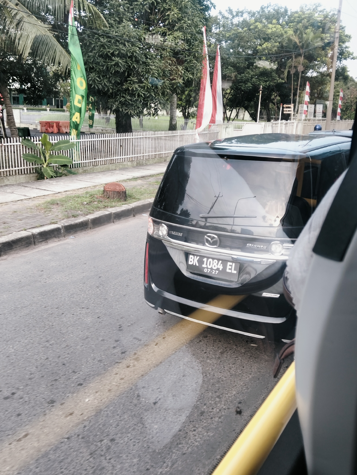 An eye-level, full shot captures a black Mazda Biante minivan on a city street in Medan, Indonesia. The minivan, angled towards the right, displays a license plate that reads "BK 1084 EL" and "07-27". The street is paved and gray, with a yellow line running along the lane. The minivan is parked near a sidewalk with a white fence. Behind the fence, a lush green tree and a banana plant are visible. In the background, other trees and buildings can be seen. Several flags are also present: a green flag with yellow lettering and two red and white flags. The overall scene suggests a bright, daytime setting.