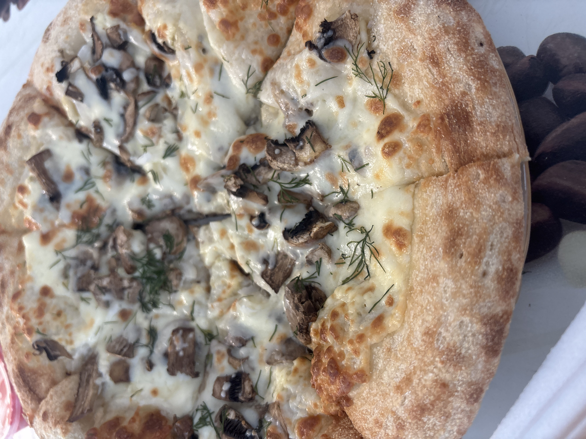 A top-down, close-up shot shows a partially eaten pizza. The pizza is cut into slices, and about half of it features visible toppings, including mushrooms and dill sprigs. The cheese appears to be melted and bubbly. The pizza crust is thick and has a slightly golden-brown color.
To the right of the pizza, there's a collection of dark brown, round objects, possibly olives or other small, dark fruits, visible through a transparent container or wrapping. A glimpse of a white napkin or paper is visible at the bottom right corner of the frame.
There is no information in this image about the city of Chaikovsky, Russia.