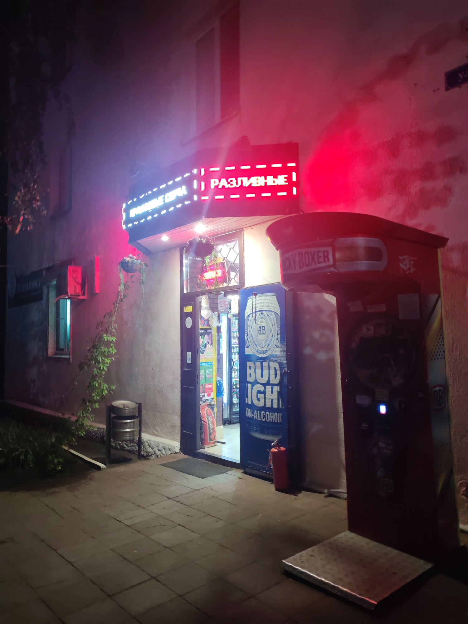 This is a nighttime exterior shot of a storefront in Vladimir, Russia. The building is a light-colored stucco, with a large, dark red boxing game machine prominently placed in front of the entrance to the right. The game machine has "LUCKY BOXER" written across the top and a digital screen at the bottom. To the left of the entrance, a large blue Bud Light non-alcoholic beer advertisement is visible. Above the entrance, a bright neon sign glows in white and pink, spelling out "РАЗЛИВНЫЕ НАПИТКИ" (meaning "Draft Drinks" or "Tap Drinks"). Above the neon sign, a red light casts a glow on the building's facade. A fire extinguisher is visible near the entrance. To the left of the doorway, a window is open, and an air conditioning unit is mounted above it. A vine with green leaves hangs down from the wall, partially obscuring a silver trash receptacle with two kegs inside. The ground is paved with gray square tiles, and there is some foliage and a drainage ditch on the left side of the image. The overall scene suggests a small shop or bar open late at night.